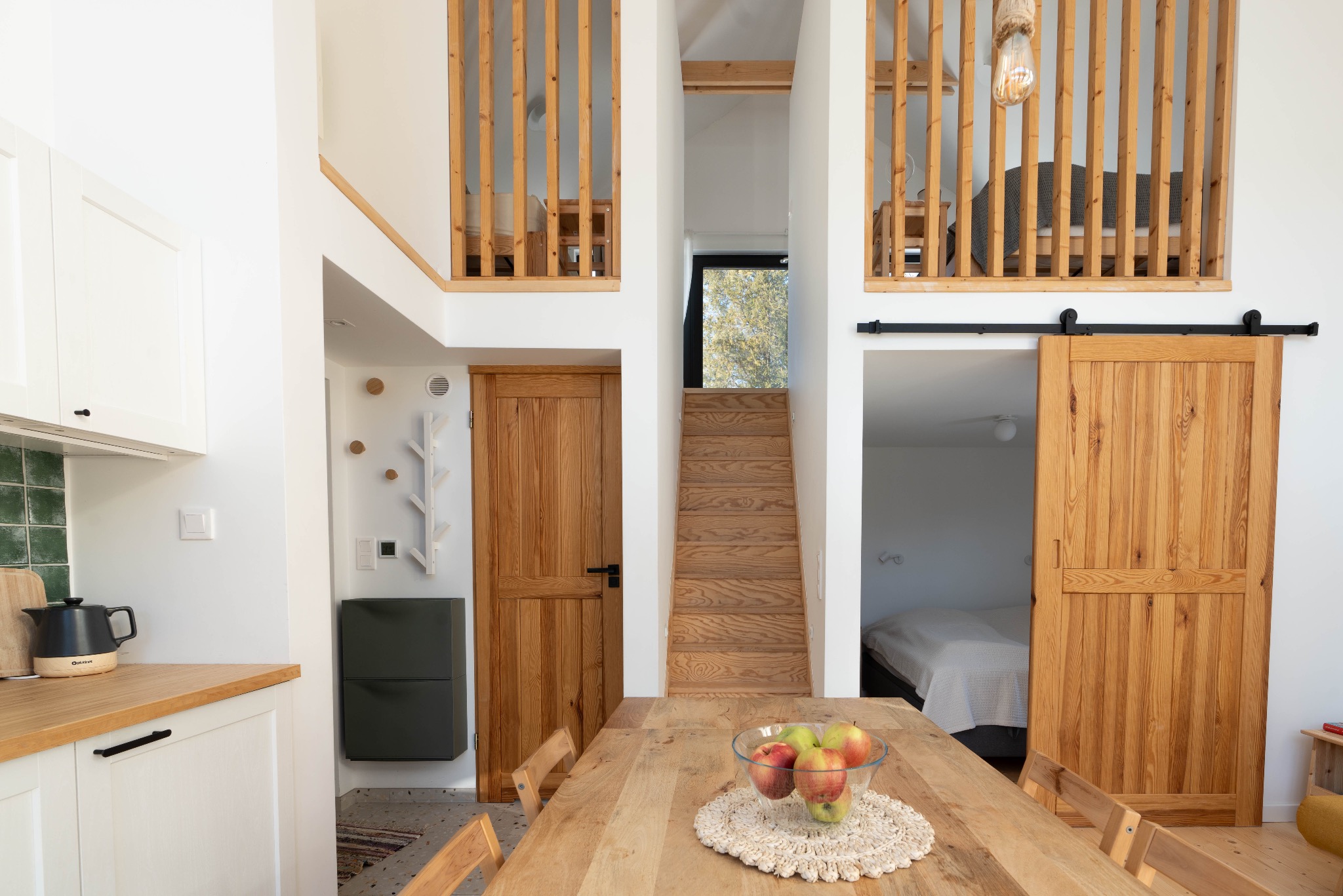 Jasne wnętrze domu z drewnianymi schodami, balustradami i przesuwanymi drzwiami stodołowymi, stół z owocami na pierwszym planie, widok z kuchni na sypialnię na antresoli.