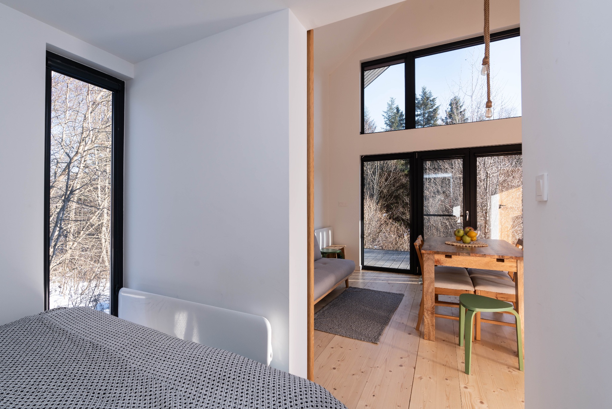 Jasne wnętrze domu z widokiem na zimowy krajobraz za oknem, drewniany stół z owocami, szara sofa i dywan, minimalistyczny design.