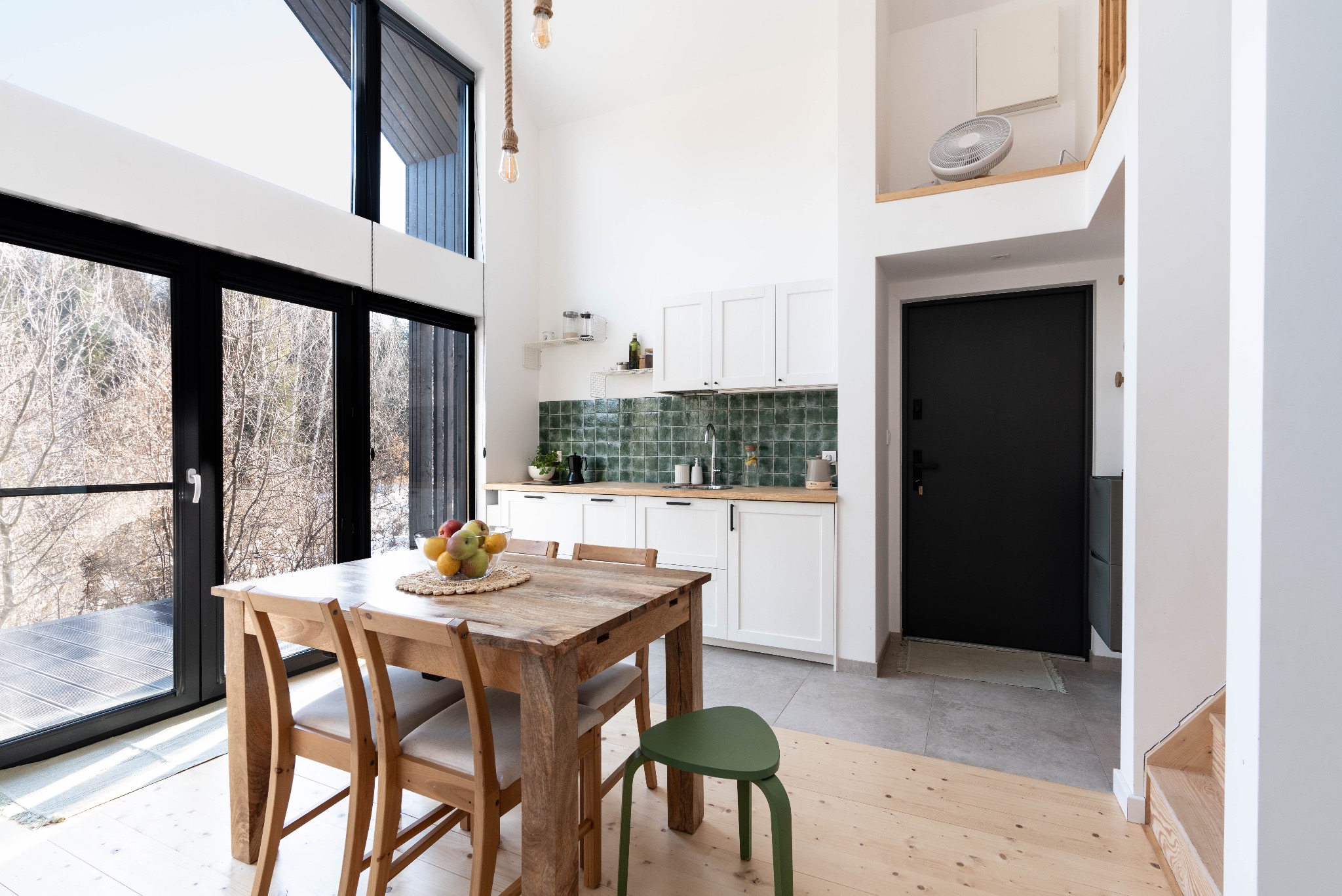 Jasna kuchnia w stylu skandynawskim z drewnianym stołem i krzesłami, owocami w misie, dużym oknem z widokiem na las i zielonymi płytkami nad blatem.