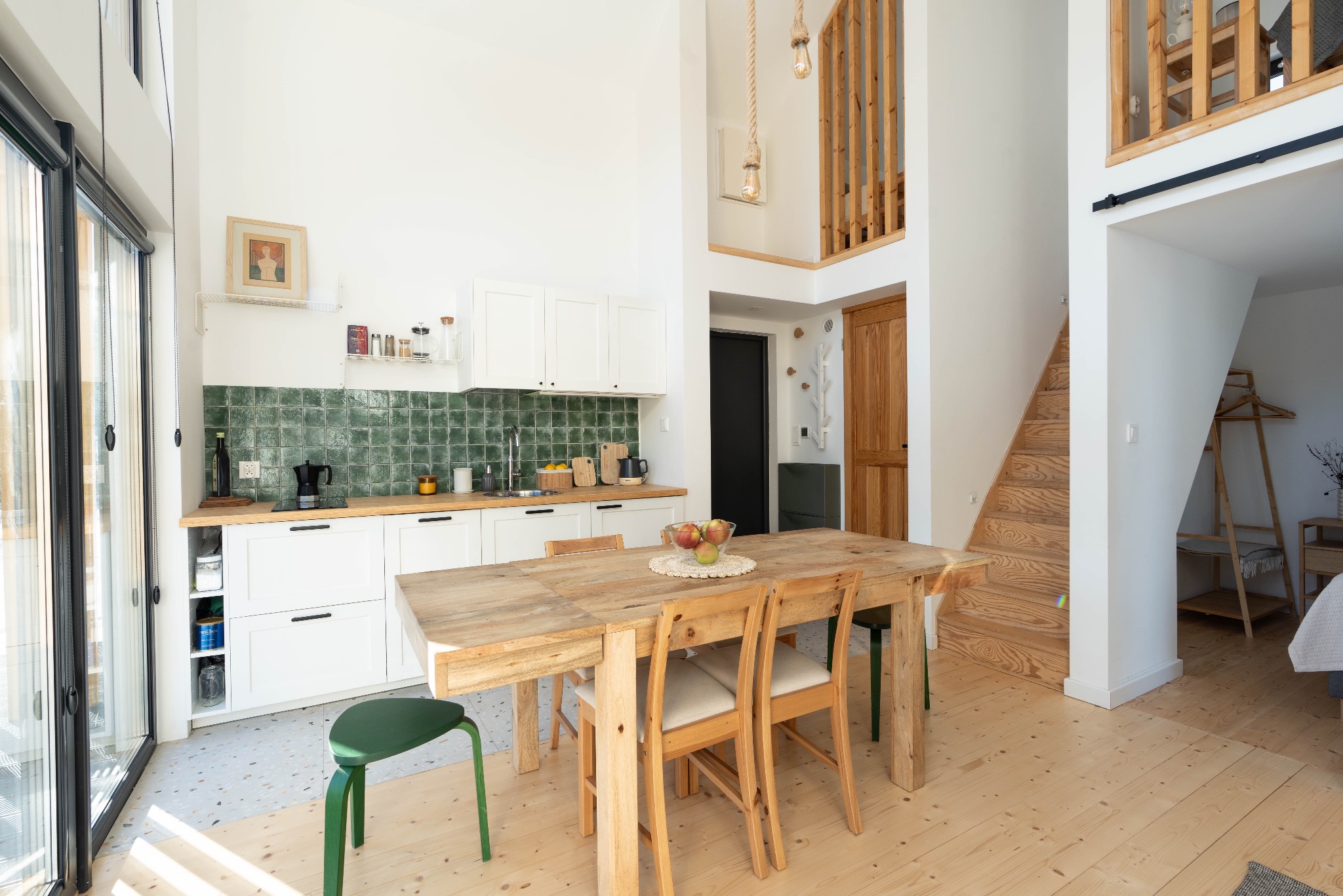 Jasne, przestronne wnętrze domu z drewnianą podłogą i stołem, zielona mozaika na ścianie w kuchni, duże okno i antresola z drewnianymi schodami.