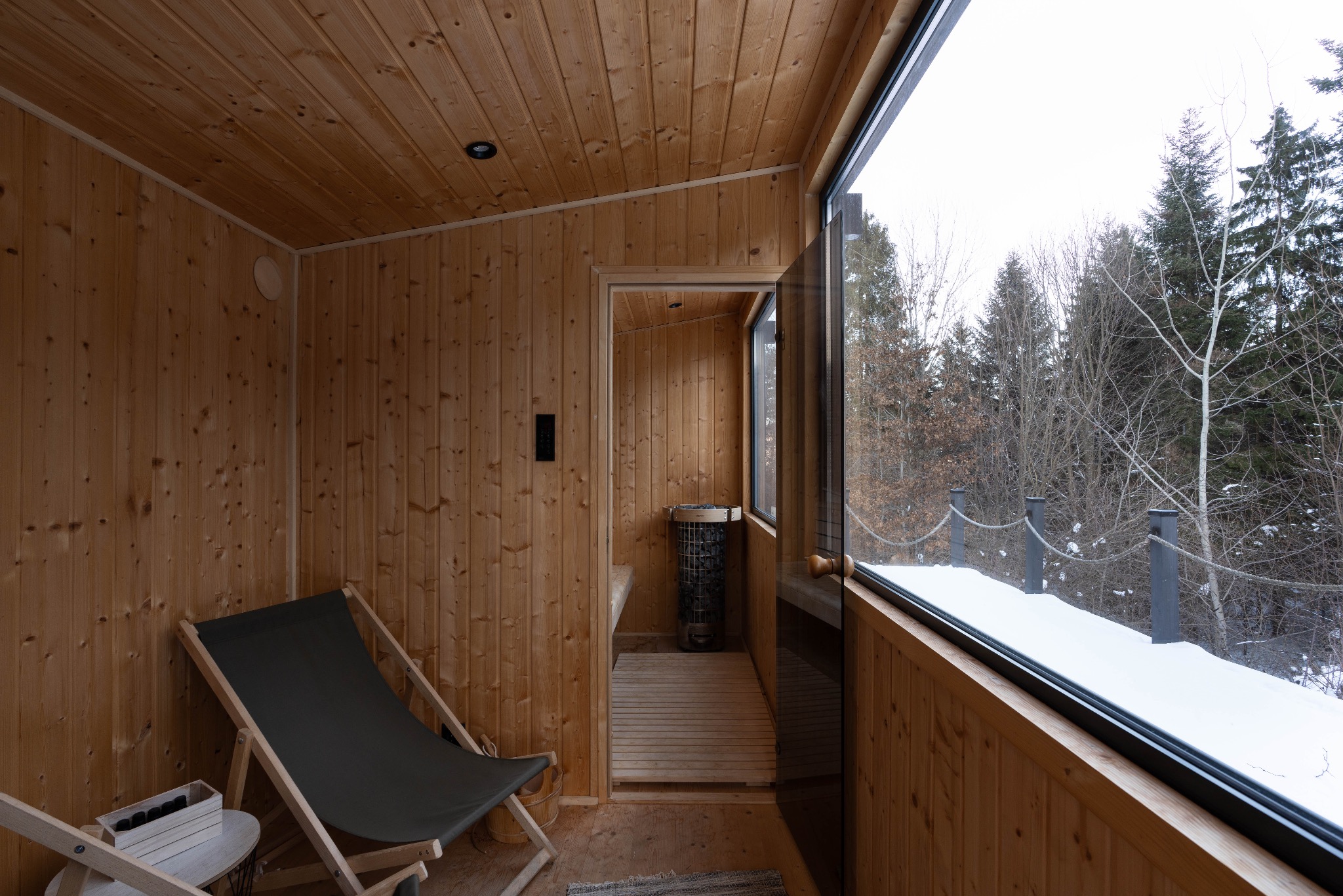 Drewniana sauna z leżakiem i widokiem na zasypany śniegiem las przez duże okno, piec do sauny widoczny w głębi.
