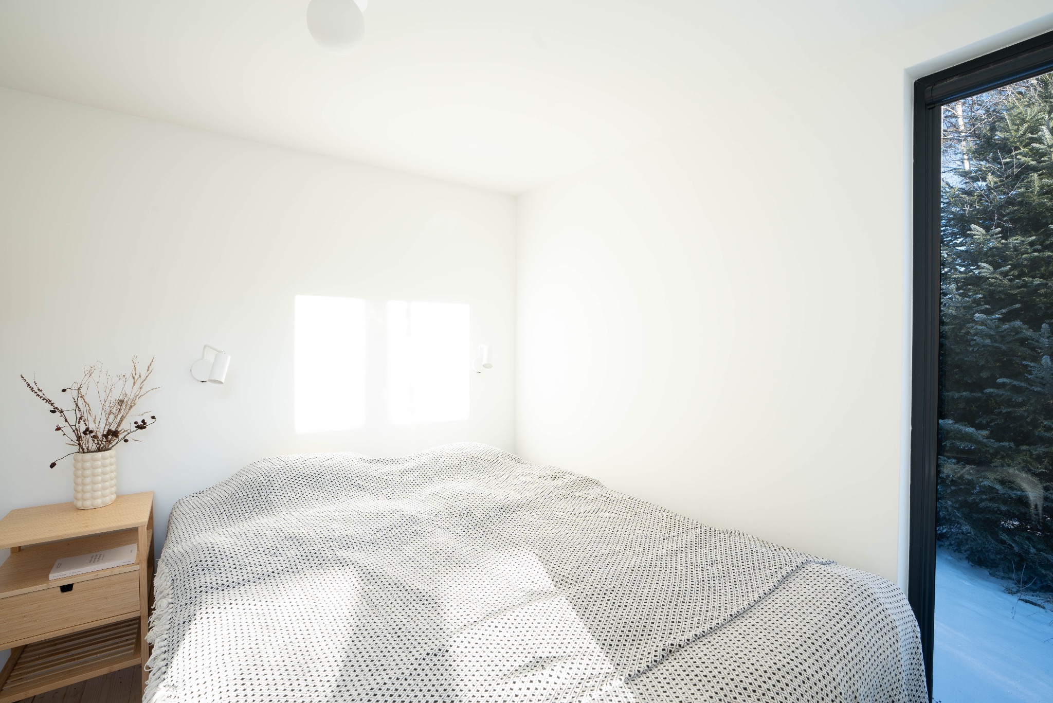 Jasna sypialnia z łóżkiem przykrytym biało-szarą narzutą w geometryczny wzór, obok drewniany stolik nocny z wazonem suchych roślin i widokiem na ośnieżone drzewa za oknem.