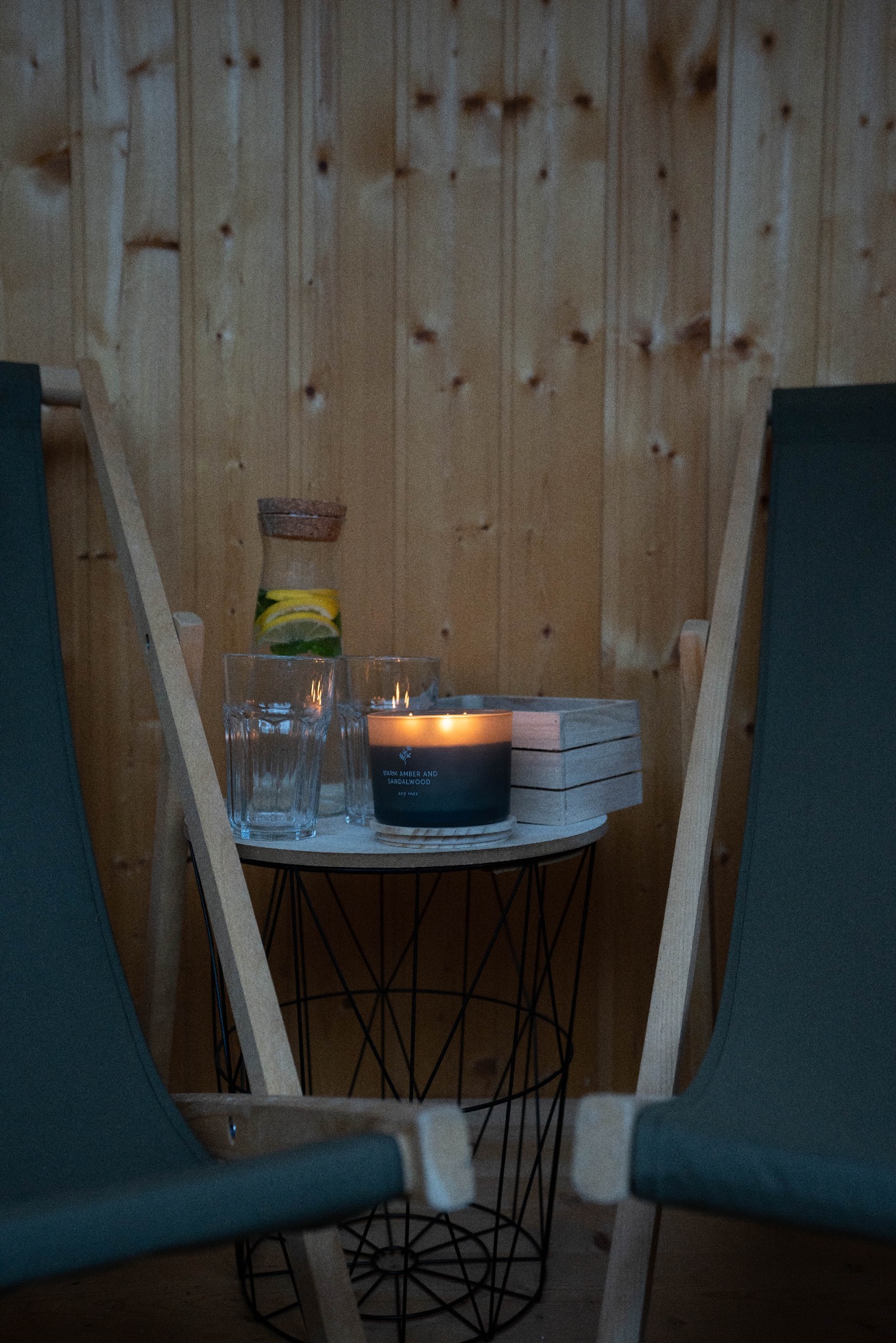 Stolik z metalowym koszem, na nim zapalona świeca Warm Amber and Sandalwood, szklanki, karafka z wodą i cytryną, drewniane podkładki, w tle drewniana ściana i fragmenty dwóch leżaków.