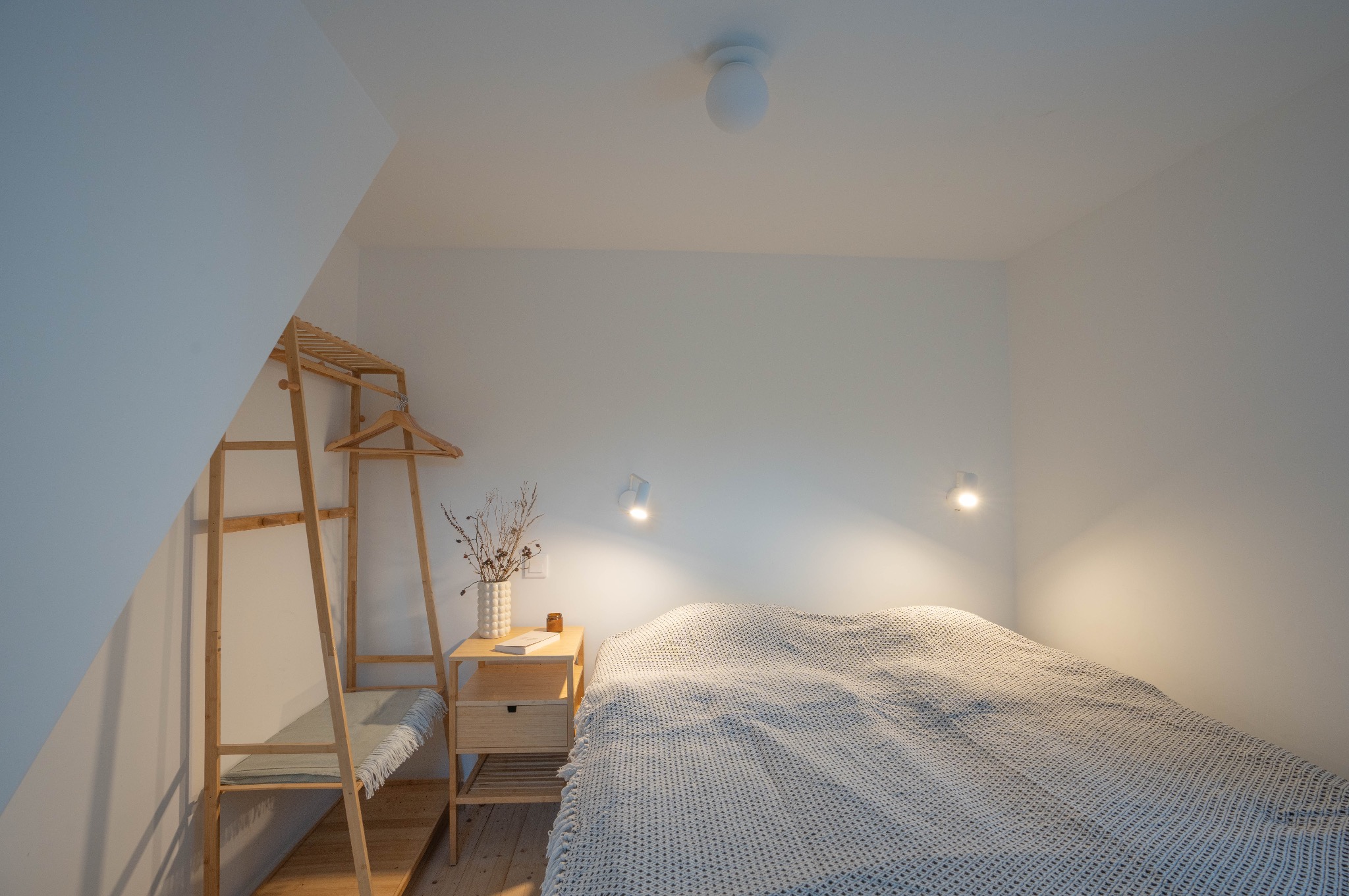 Jasna sypialnia w stylu skandynawskim z drewnianym regałem na ubrania, stolikiem nocnym z wazonem z suszonymi kwiatami i książką, oraz łóżkiem przykrytym szarą narzutą w gruby splot.