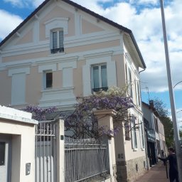 Kambud - Dwupiętrowy dom z różową elewacją i białymi zdobieniami, częściowo przysłonięty kwitnącą glicynią i metalowym ogrodzeniem, widziany z perspektywy ulicy w słoneczny dzień.