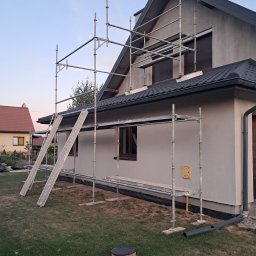 Kambud - Dom jednorodzinny w trakcie prac elewacyjnych, widoczne rusztowanie, świeżo otynkowana szara ściana, czarna dachówka, żółta skrzynka gazowa na elewacji.