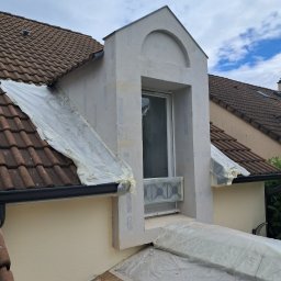 Kambud - Elewacja domu jednorodzinnego z lukarną, w trakcie prac przygotowawczych, zabezpieczona folią malarską, widoczna struktura tynku.