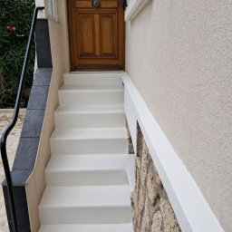 Kambud - Wejście do domu z drewnianymi drzwiami po odnowieniu, schody pomalowane na biało, elewacja w odcieniach beżu z dekoracyjnym kamieniem.