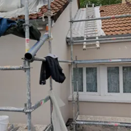 Świeżo otynkowana elewacja domu jednorodzinnego w kolorze beżowym, widok z rusztowania, zabezpieczona folią okienna stolarka, dach z czerwonej dachówki, drabina oparta o dach.