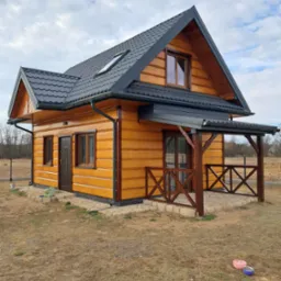 Nowoczesny, parterowy dom z drewnianą elewacją w odcieniach miodowego brązu, ciemnoszary dach z oknem połaciowym, zadaszony taras z drewnianą balustradą, widoczne miski dla zwierząt na trawniku.