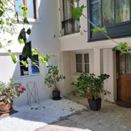 Biała elewacja budynku z oknami i drewnianymi drzwiami, dekorowana roślinami w donicach i białymi kamykami, widziana przez liście drzewa.