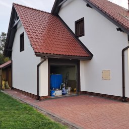 Kambud - Biała elewacja domu z brązowymi detalami, widok na otwarty garaż z narzędziami malarskimi. Czerwona dachówka i kostka brukowa. Świeżo pomalowana ściana.