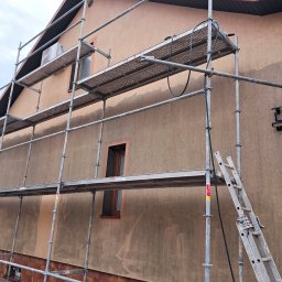 Kambud - Dom w trakcie prac elewacyjnych: rusztowanie przy ścianie z widocznymi śladami po myciu. Fragment dachu z brązową dachówką. Widoczne okna i klimatyzator.