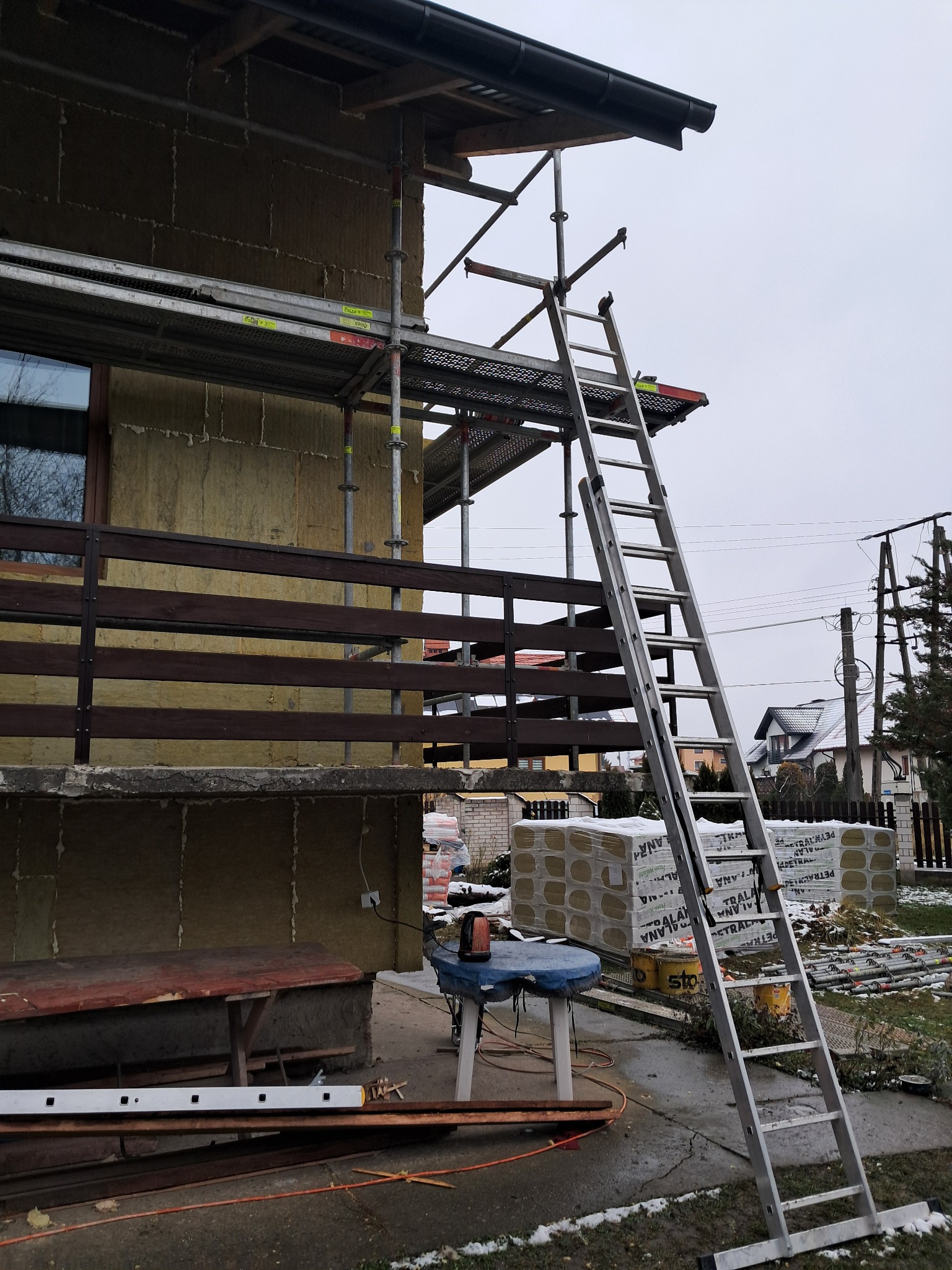 Dom w trakcie prac z użyciem rusztowania i drabiny, elewacja pokryta warstwą izolacyjną, widoczne paczki materiału izolacyjnego na zewnątrz.