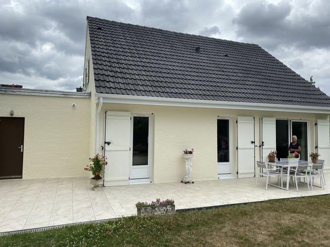 Jasna elewacja domu jednorodzinnego z ciemnym dachem, białymi okiennicami i tarasem wyłożonym płytkami, widok z ogrodu w pochmurny dzień.