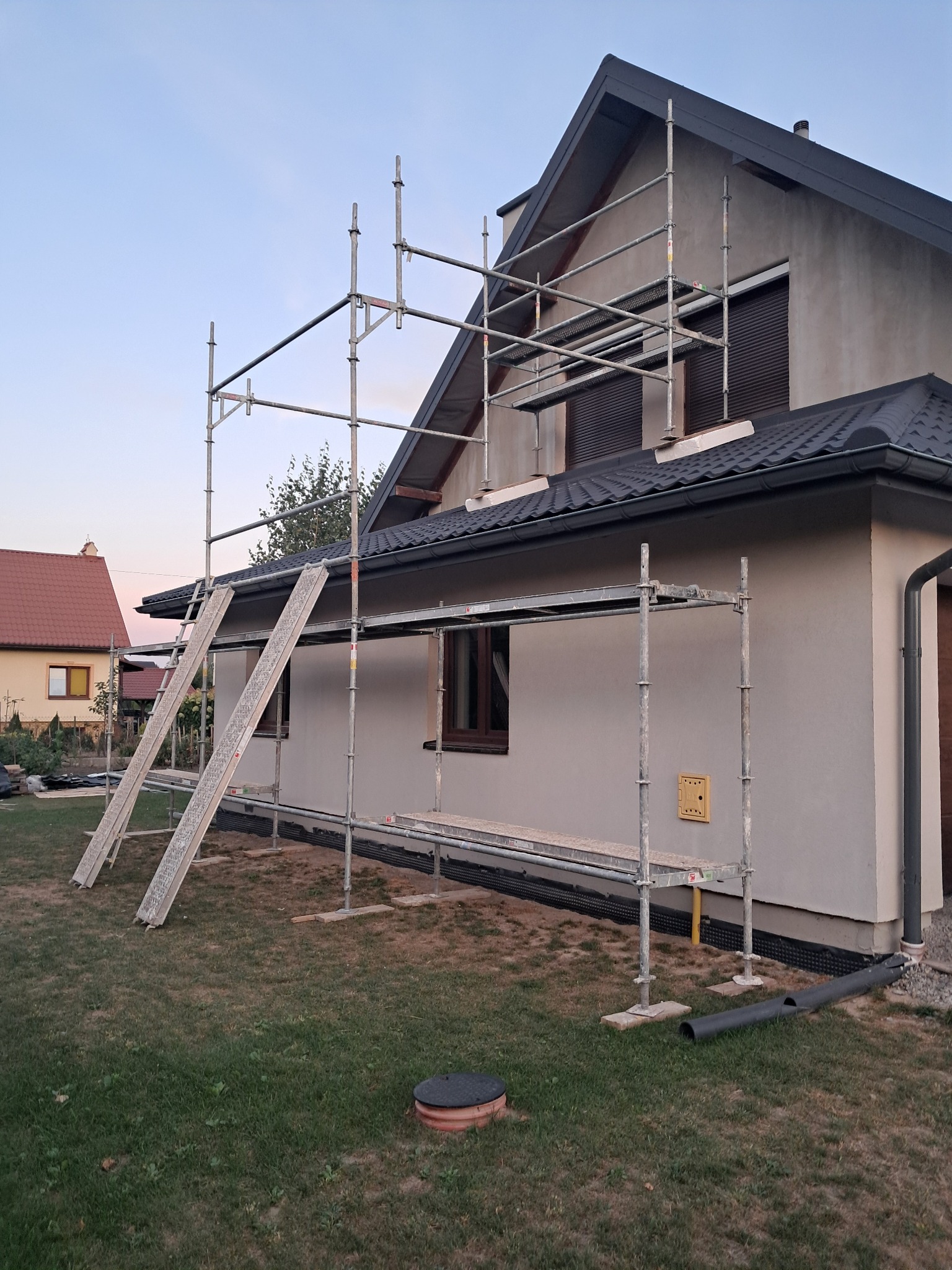 Dom jednorodzinny w trakcie prac elewacyjnych, widoczne rusztowania, szara elewacja w stanie surowym, czarny dach i okna z roletami zewnętrznymi.