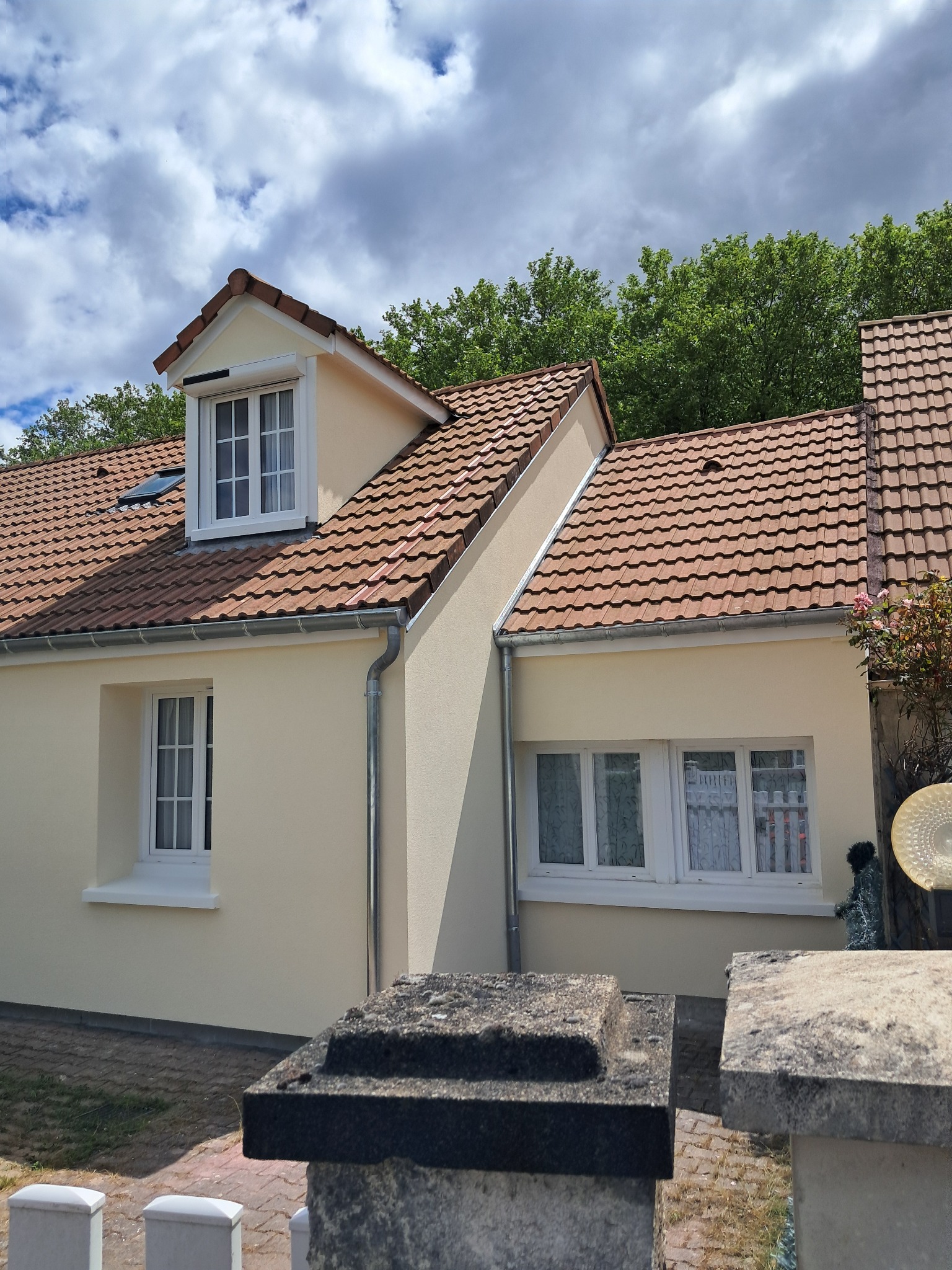 Jasnożółty dom jednorodzinny z czerwoną dachówką i białymi oknami, widoczny zza kamiennego murku.