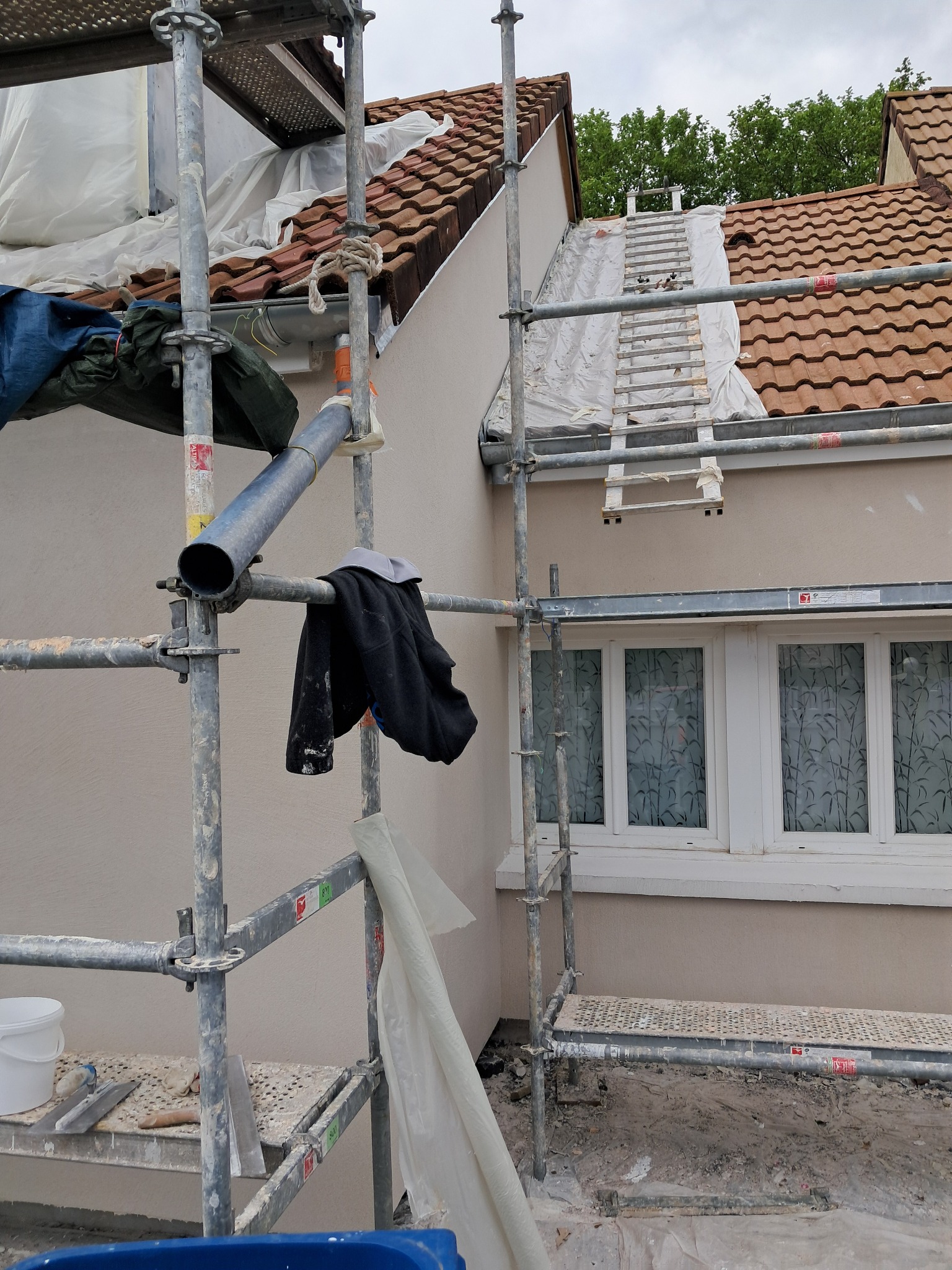 Świeżo otynkowana elewacja domu jednorodzinnego w kolorze beżowym, widok z rusztowania, zabezpieczona folią okienna stolarka, dach z czerwonej dachówki, drabina oparta o dach.