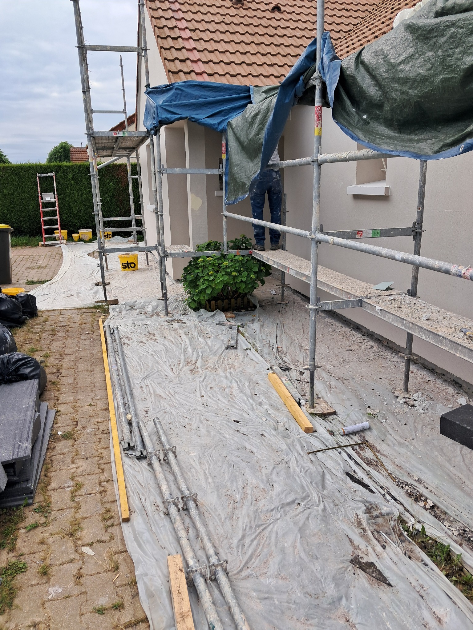 Dom jednorodzinny w trakcie prac elewacyjnych, widoczne rusztowania, zabezpieczenia folią i osoba na platformie roboczej.