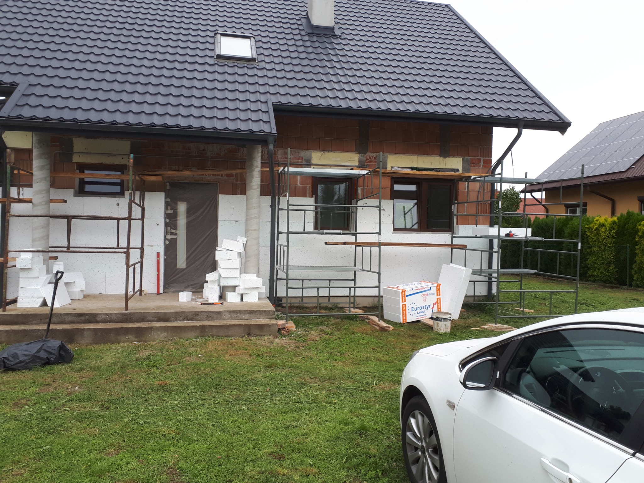 Dom w trakcie prac izolacyjnych: widoczne rusztowania, styropianowe płyty i częściowo ocieplona ściana z cegły.