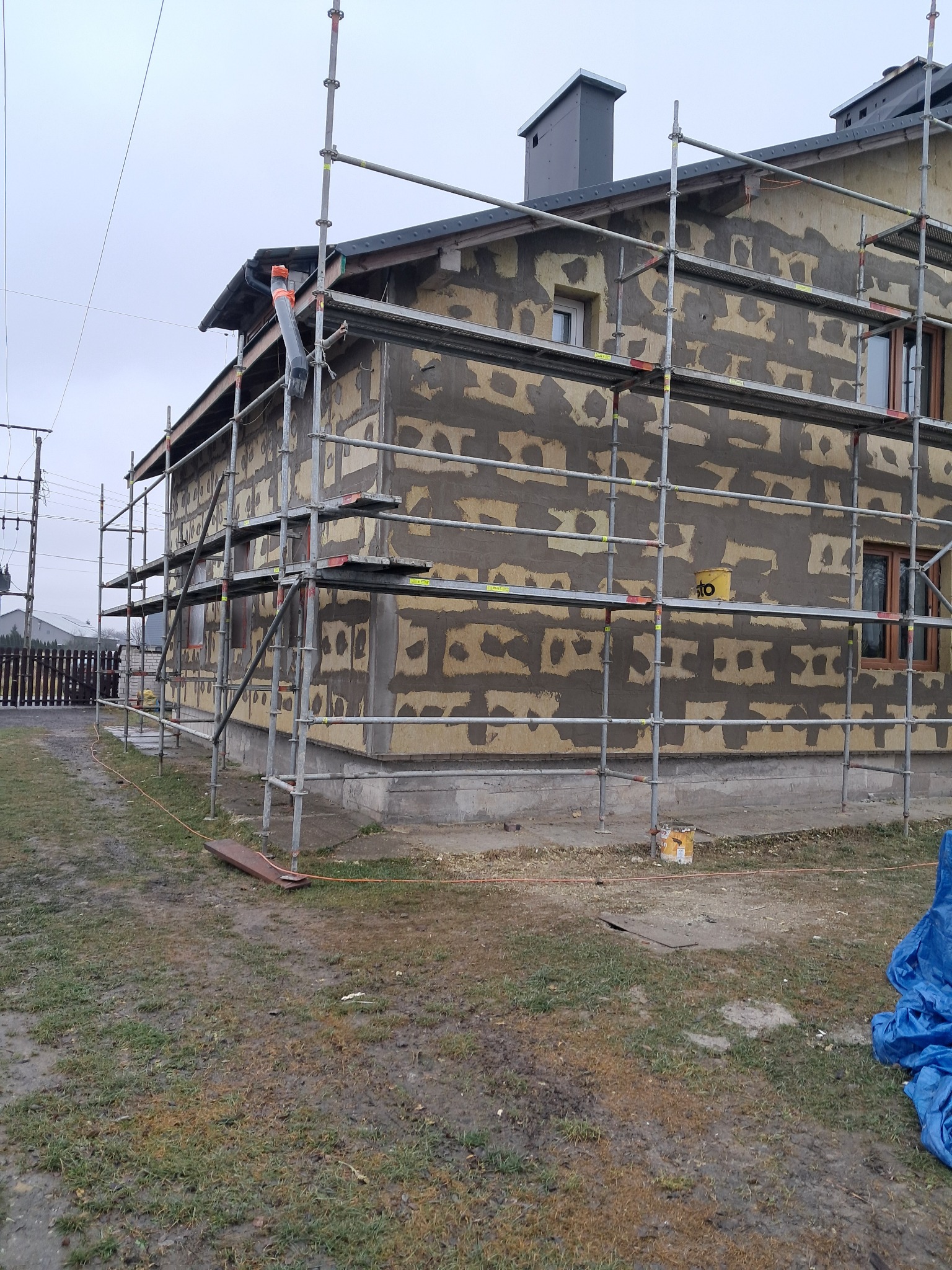 Dom jednorodzinny w trakcie przygotowania do nałożenia warstwy izolacyjnej, widoczne rusztowania, fragment ocieplenia i warstwa kleju na fasadzie, przygotowany teren wokół budynku.