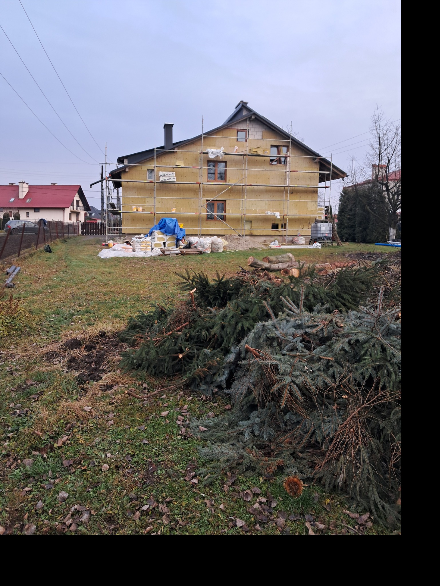 Dwupiętrowy dom w trakcie ocieplania elewacji, widoczne rusztowania, materiały izolacyjne, oraz sterta gałęzi na pierwszym planie.