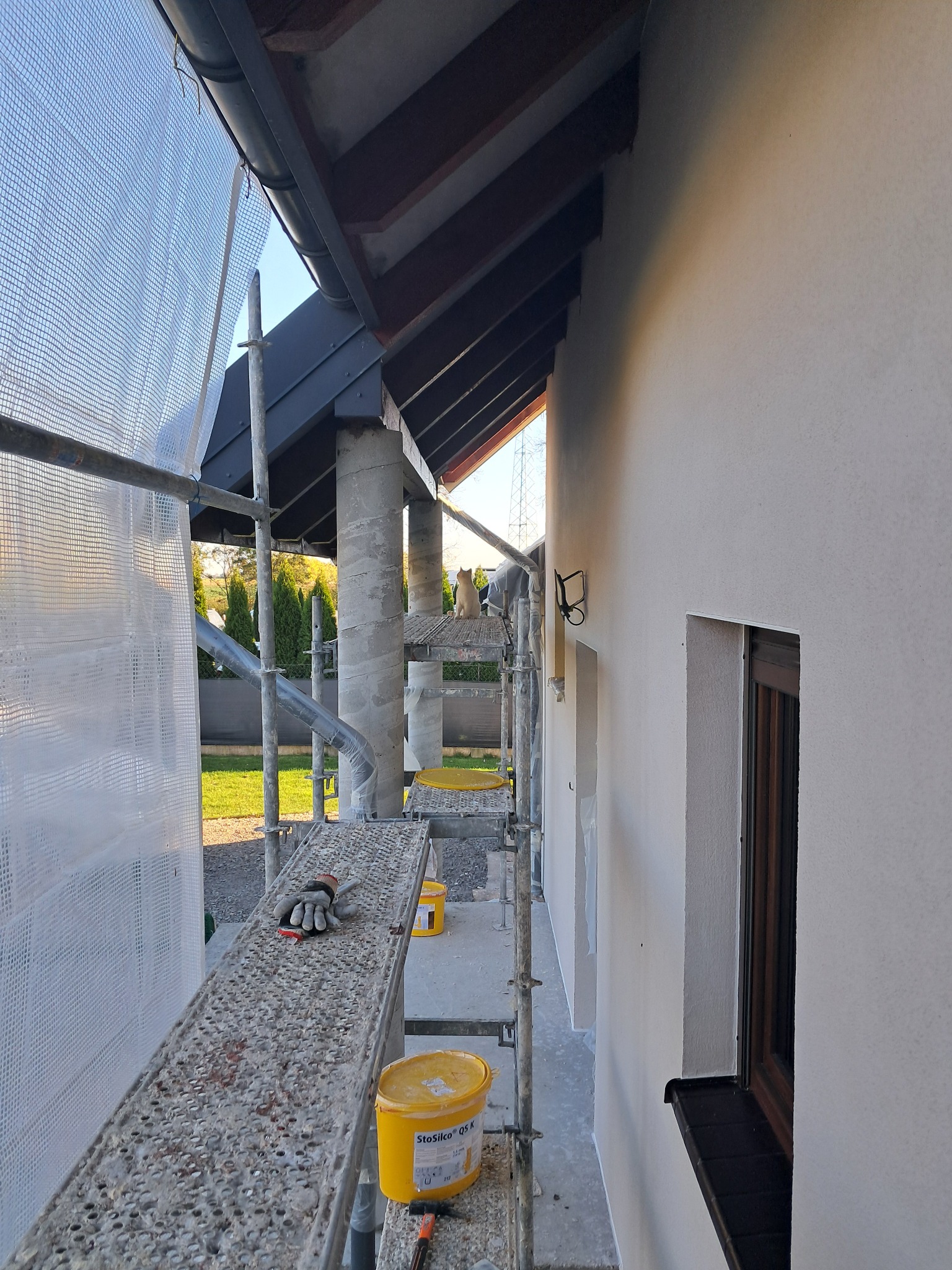 Ściana budynku w trakcie prac elewacyjnych, widoczne rusztowanie z wiaderkami z zaprawą, rękawice robocze i kot na rusztowaniu