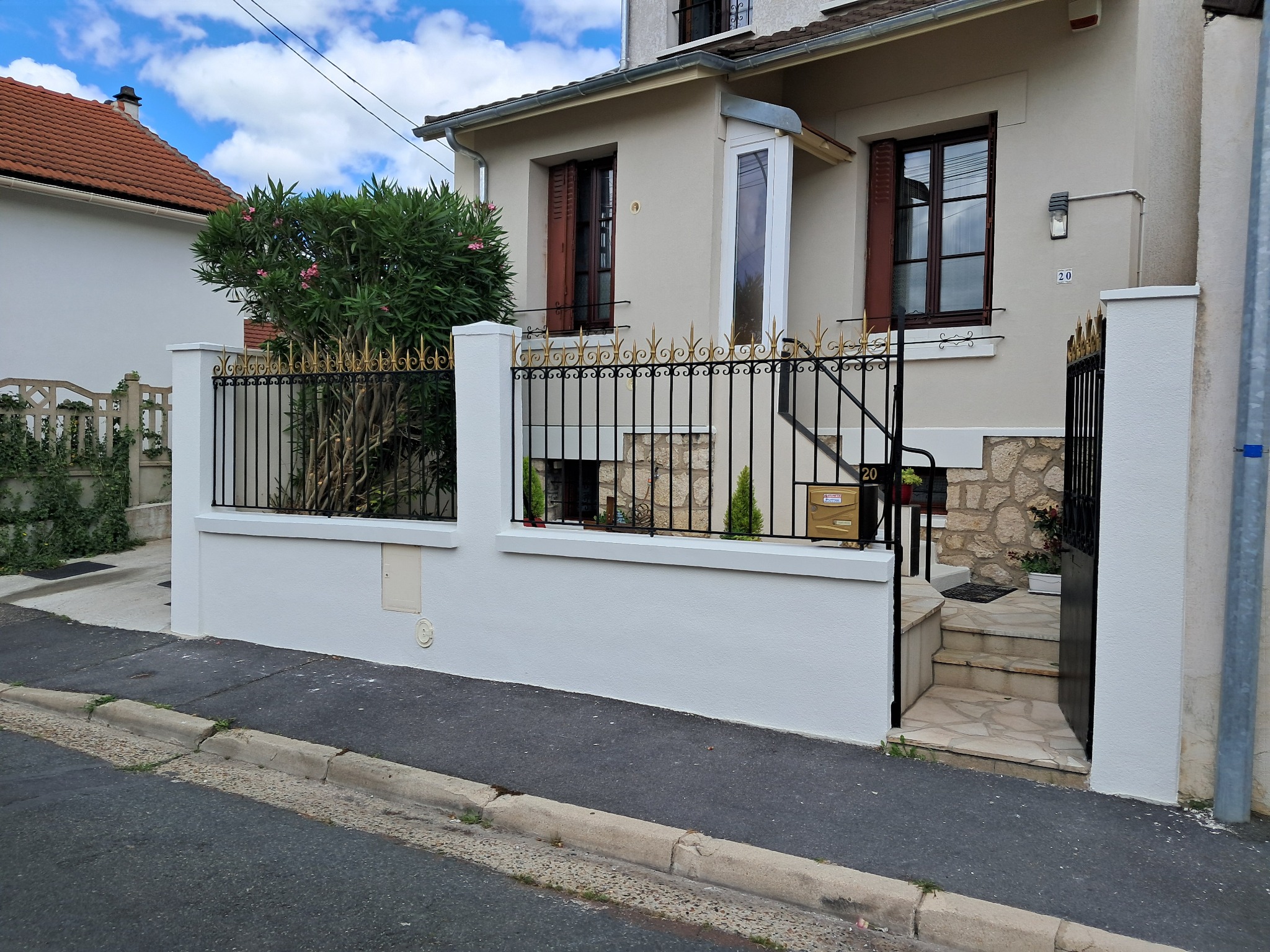 Jasna elewacja domu jednorodzinnego z dekoracyjnym, czarnym ogrodzeniem z elementami w kolorze złota, widoczne okna z brązowymi okiennicami i numer domu '20'.