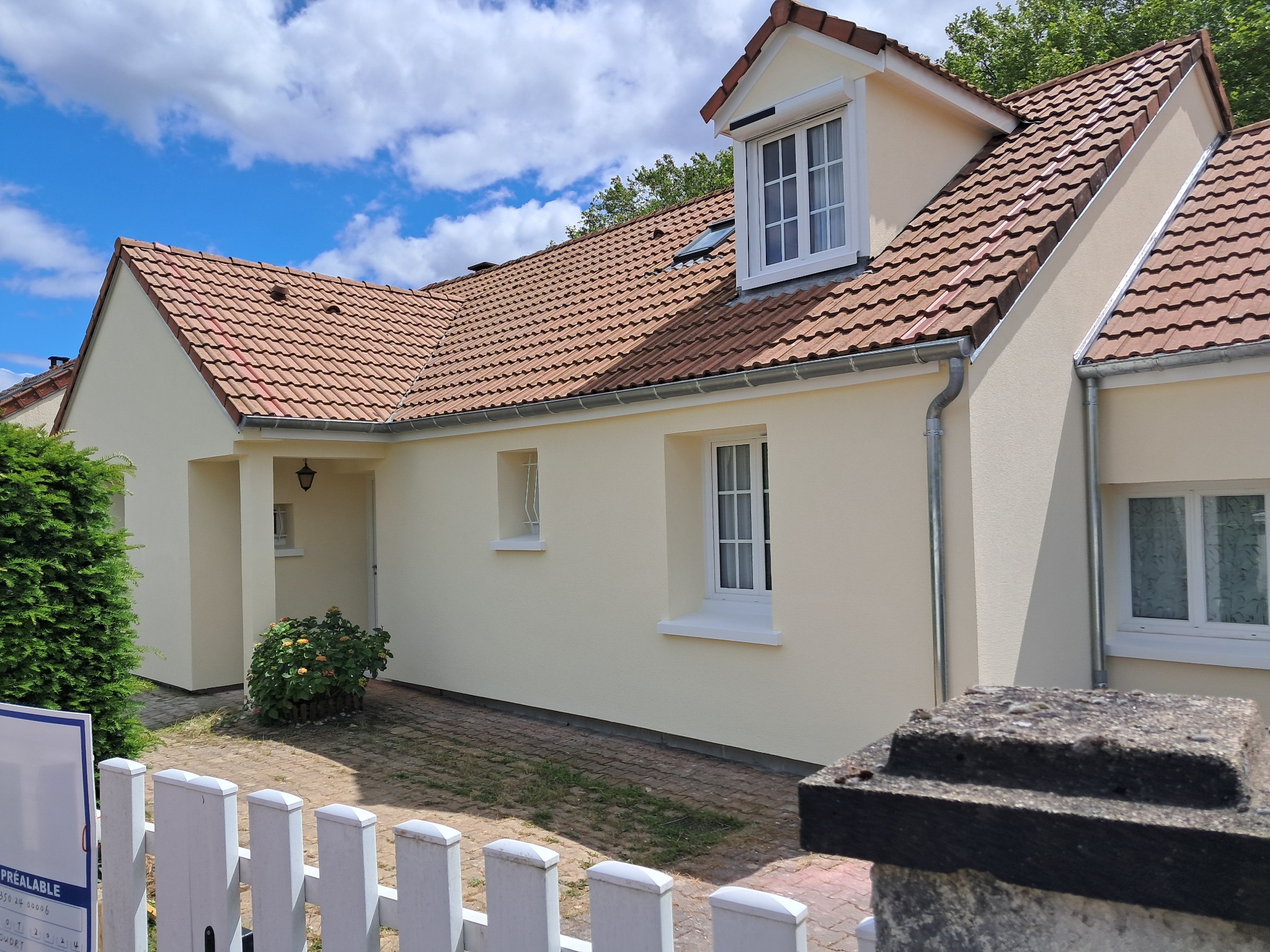 Jasna elewacja domu jednorodzinnego z brązową dachówką, widok z poziomu gruntu przez biały płotek.