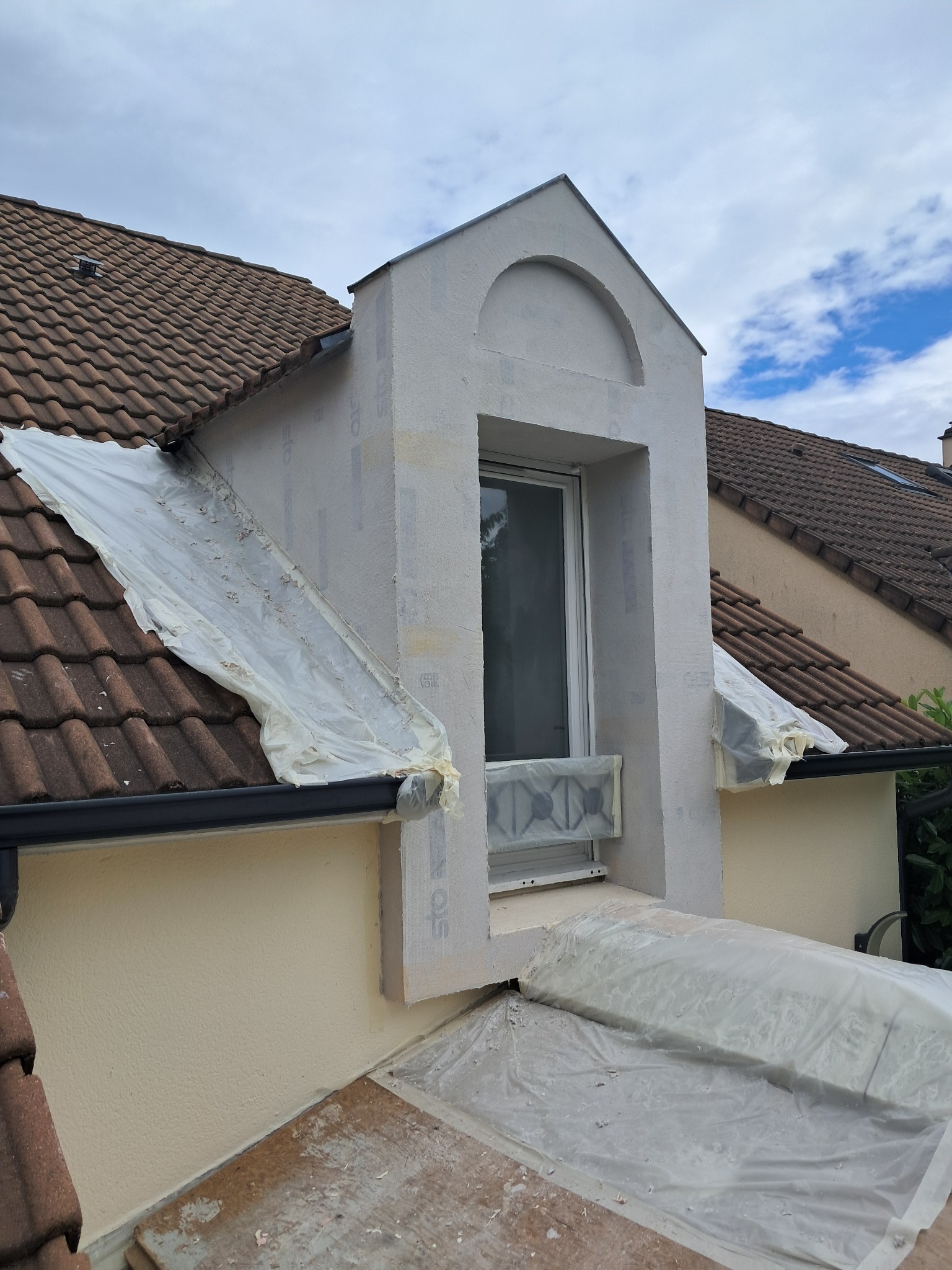 Elewacja domu w trakcie prac wykończeniowych; widoczna lukarna z oknem zabezpieczona folią, fragment dachu z dachówką ceramiczną oraz fragment otynkowanej ściany.