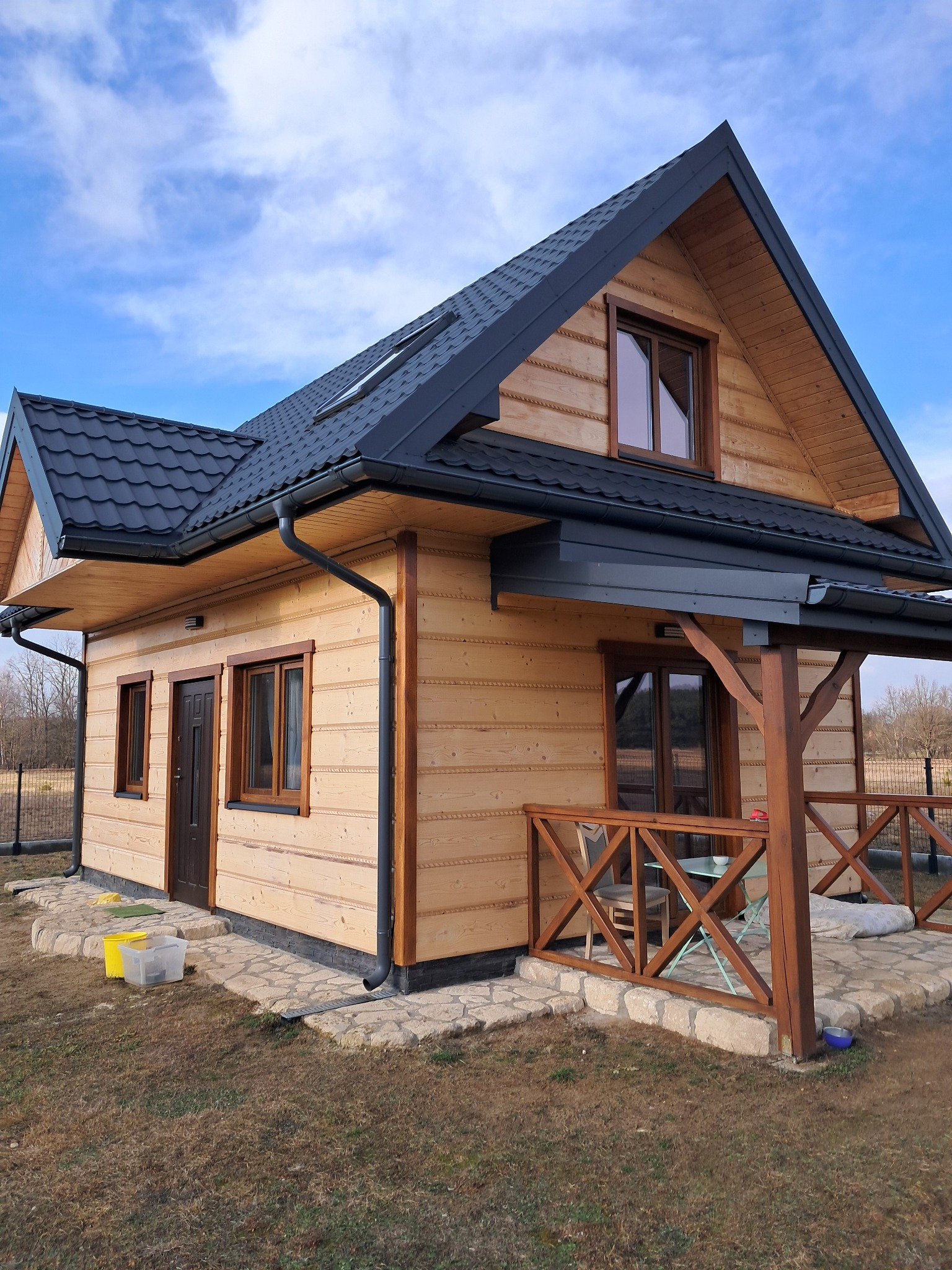 Nowoczesny dom z bali z ciemną dachówką i drewnianą werandą, widok z boku na tle błękitnego nieba z chmurami.