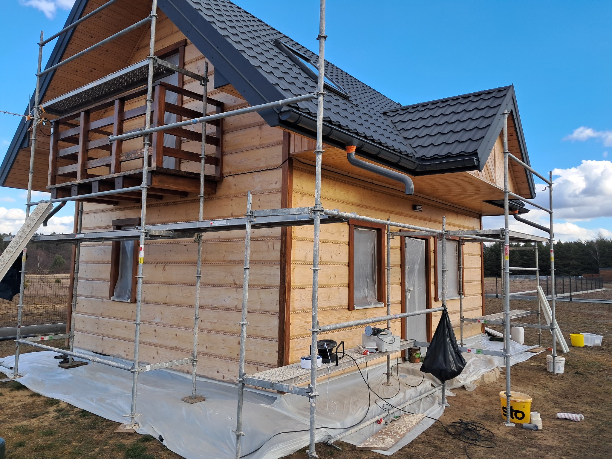 Drewniany dom w trakcie prac elewacyjnych, otoczony rusztowaniem, z widocznymi detalami wykończenia i przygotowanymi narzędziami malarskimi.