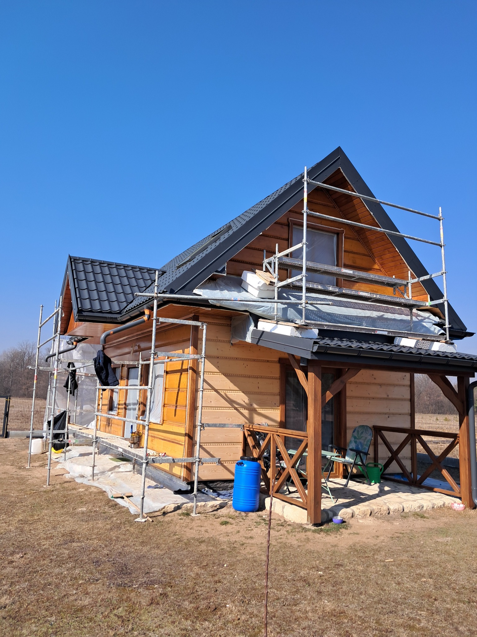 Drewniany dom w trakcie prac, rusztowania wokół, folia ochronna na oknach, widoczne materiały izolacyjne na dachu, niebieski pojemnik przy tarasie.