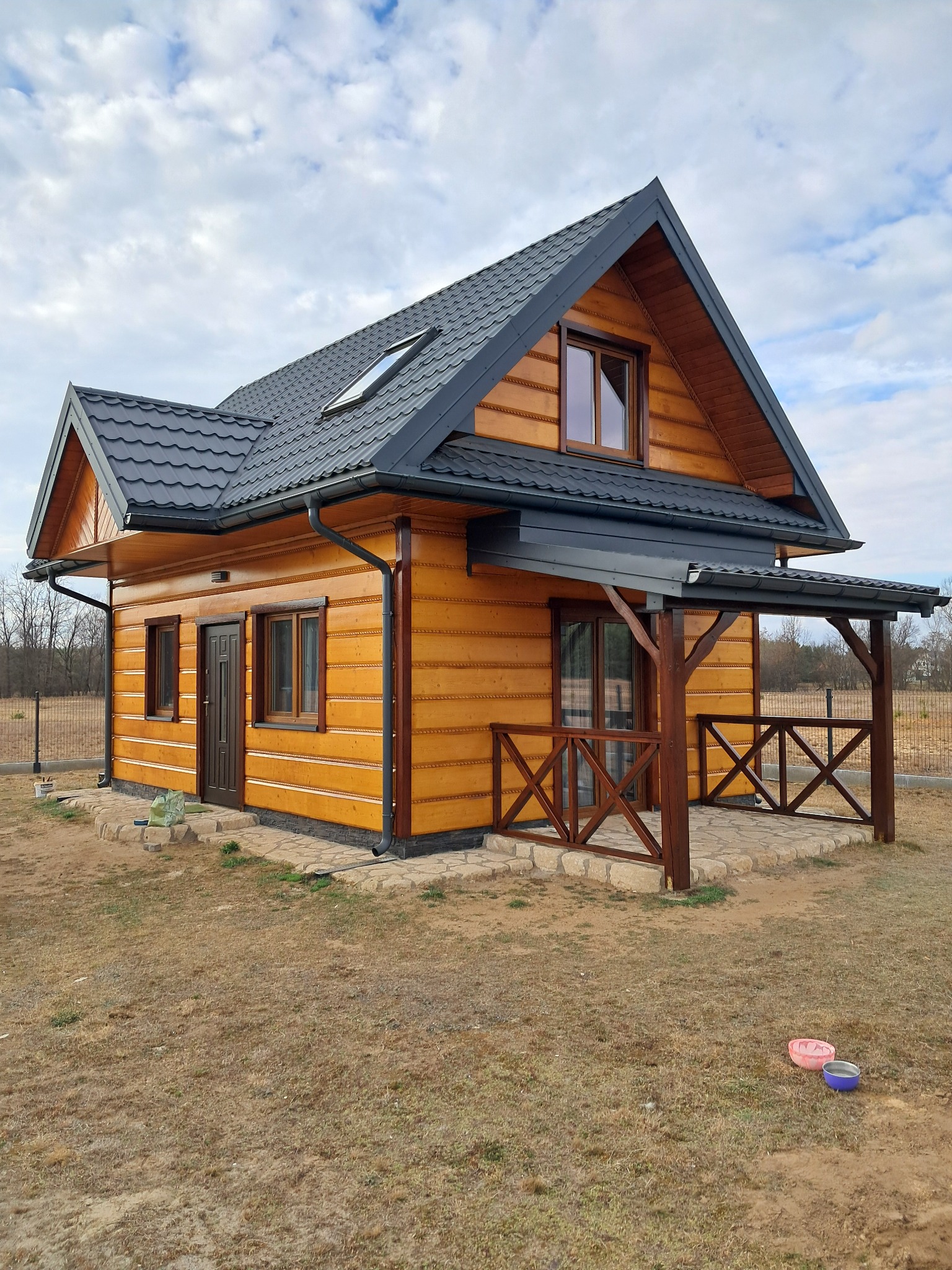 Nowoczesny, parterowy dom z drewnianą elewacją w odcieniach miodowego brązu, ciemnoszary dach z oknem połaciowym, zadaszony taras z drewnianą balustradą, widoczne miski dla zwierząt na trawniku.