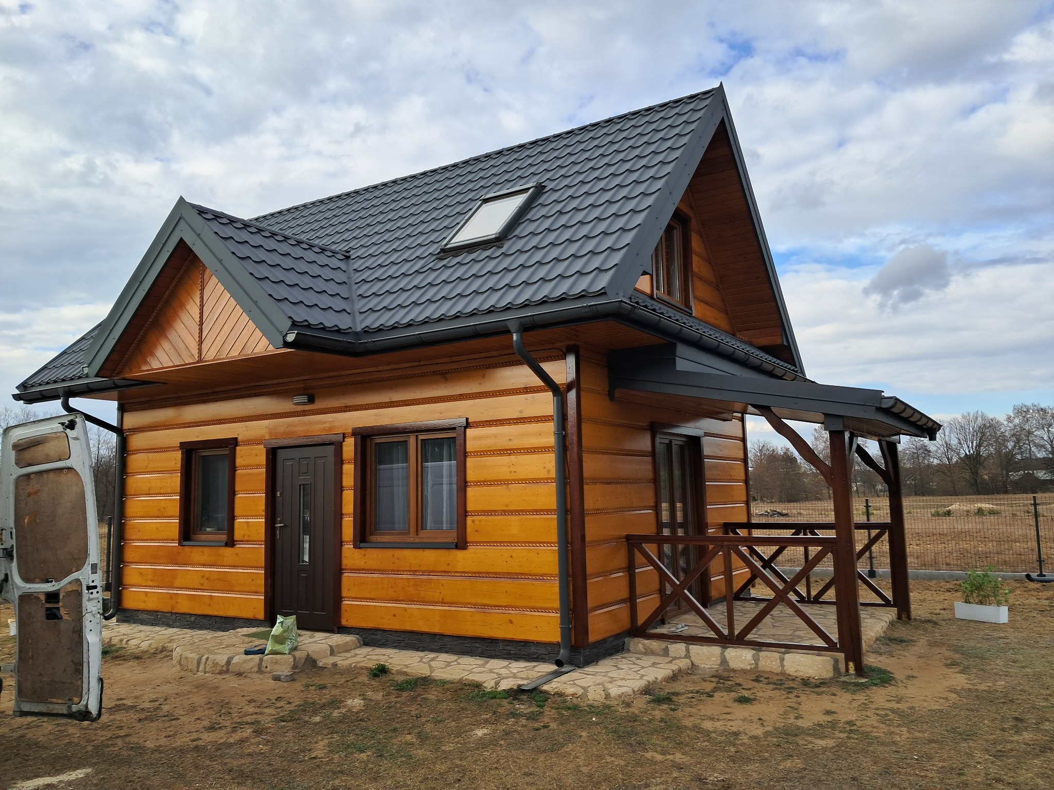 Drewniany dom z ciemnoszarą blachodachówką, widoczne detale elewacji z jasnego drewna i otwarte drzwi białego busa w tle.