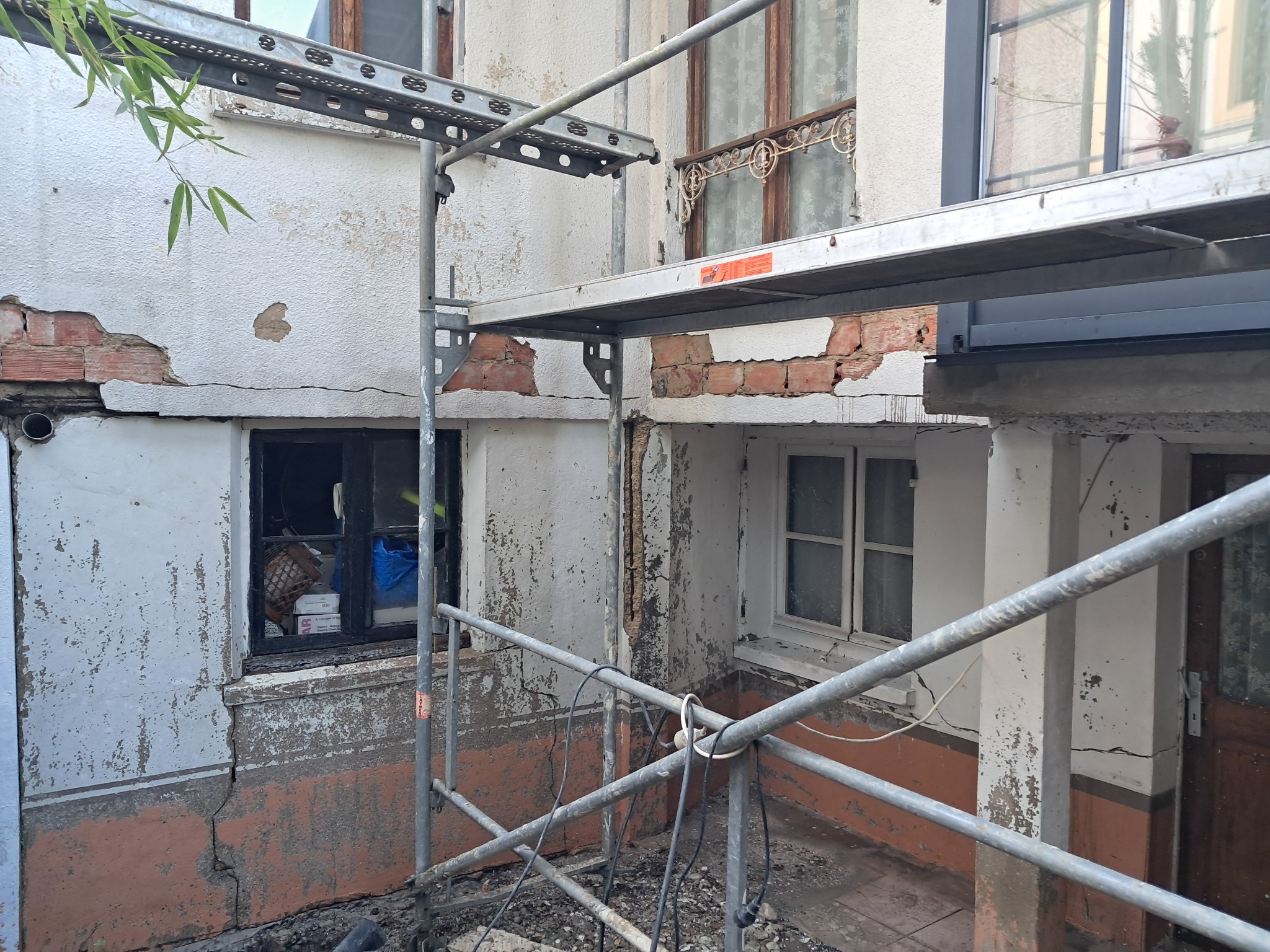 Elewacja budynku w trakcie renowacji, widoczne uszkodzenia muru i rusztowanie. Prace przygotowawcze do naprawy elewacji, widoczne fragmenty odsłoniętej cegły.