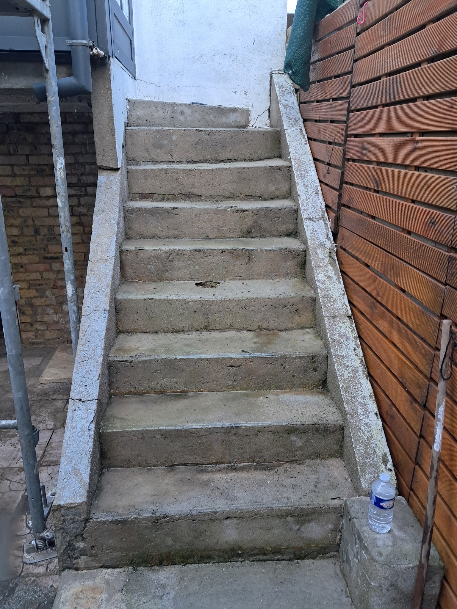 Betonowe schody zewnętrzne przed budynkiem, z widocznymi śladami użytkowania i butelką wody na dole, otoczone drewnianą ścianą i rusztowaniem.