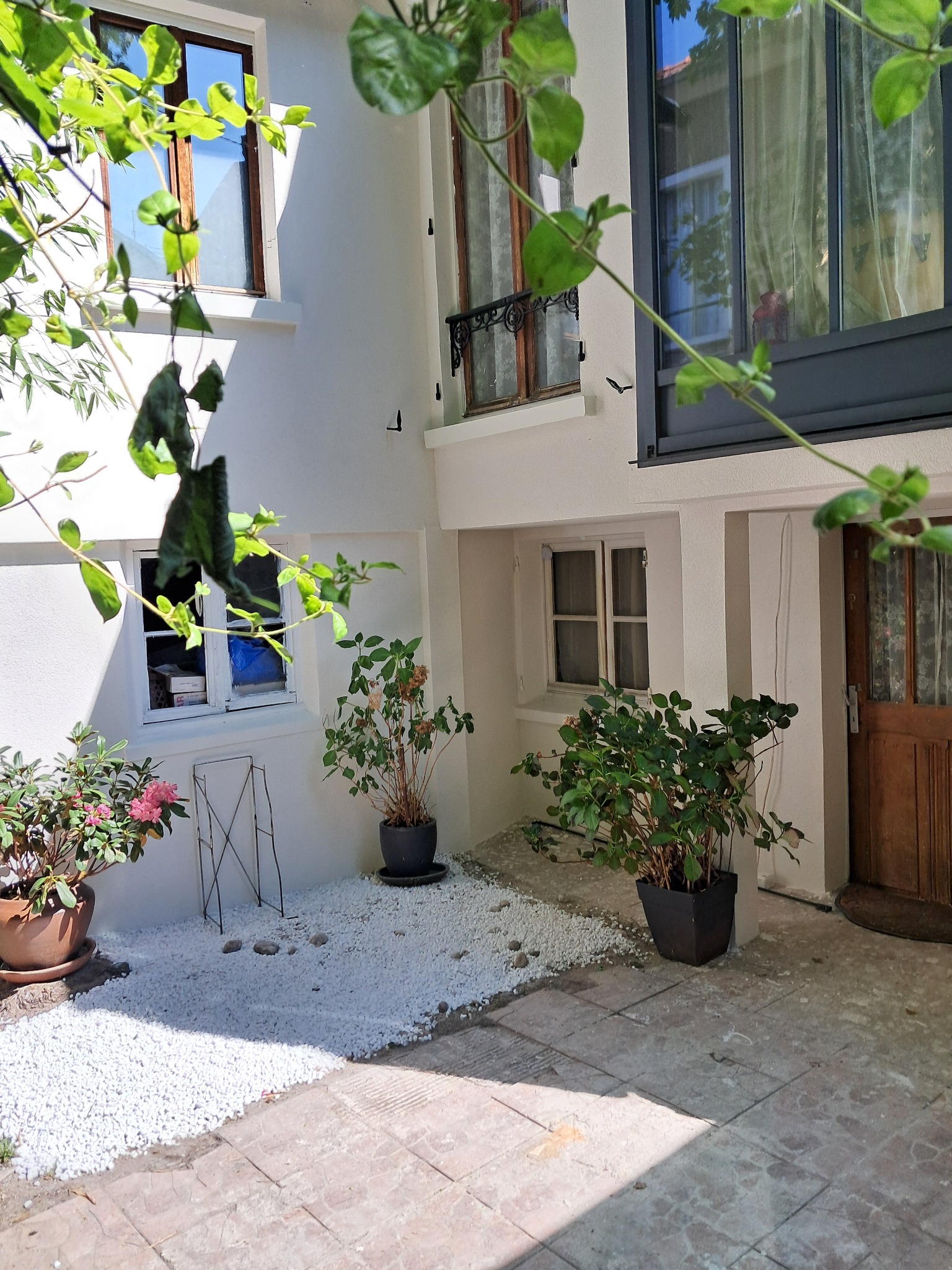 Biała elewacja budynku z oknami i drewnianymi drzwiami, dekorowana roślinami w donicach i białymi kamykami, widziana przez liście drzewa.