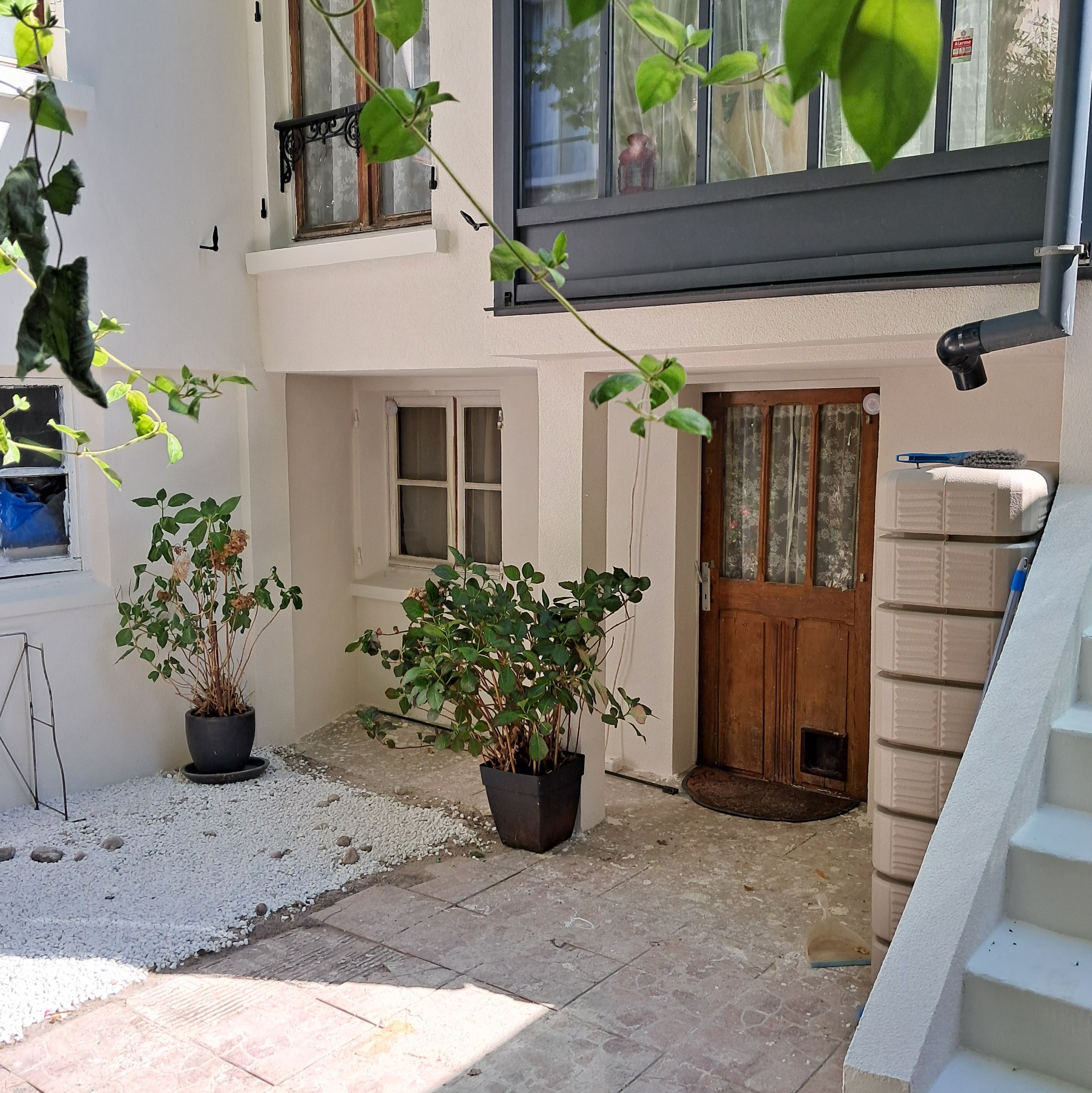 Dziedziniec po renowacji z białą elewacją, roślinami w donicach i dekoracyjnym żwirem. Widoczne narzędzia i pojemniki, sugerujące prace wykończeniowe.