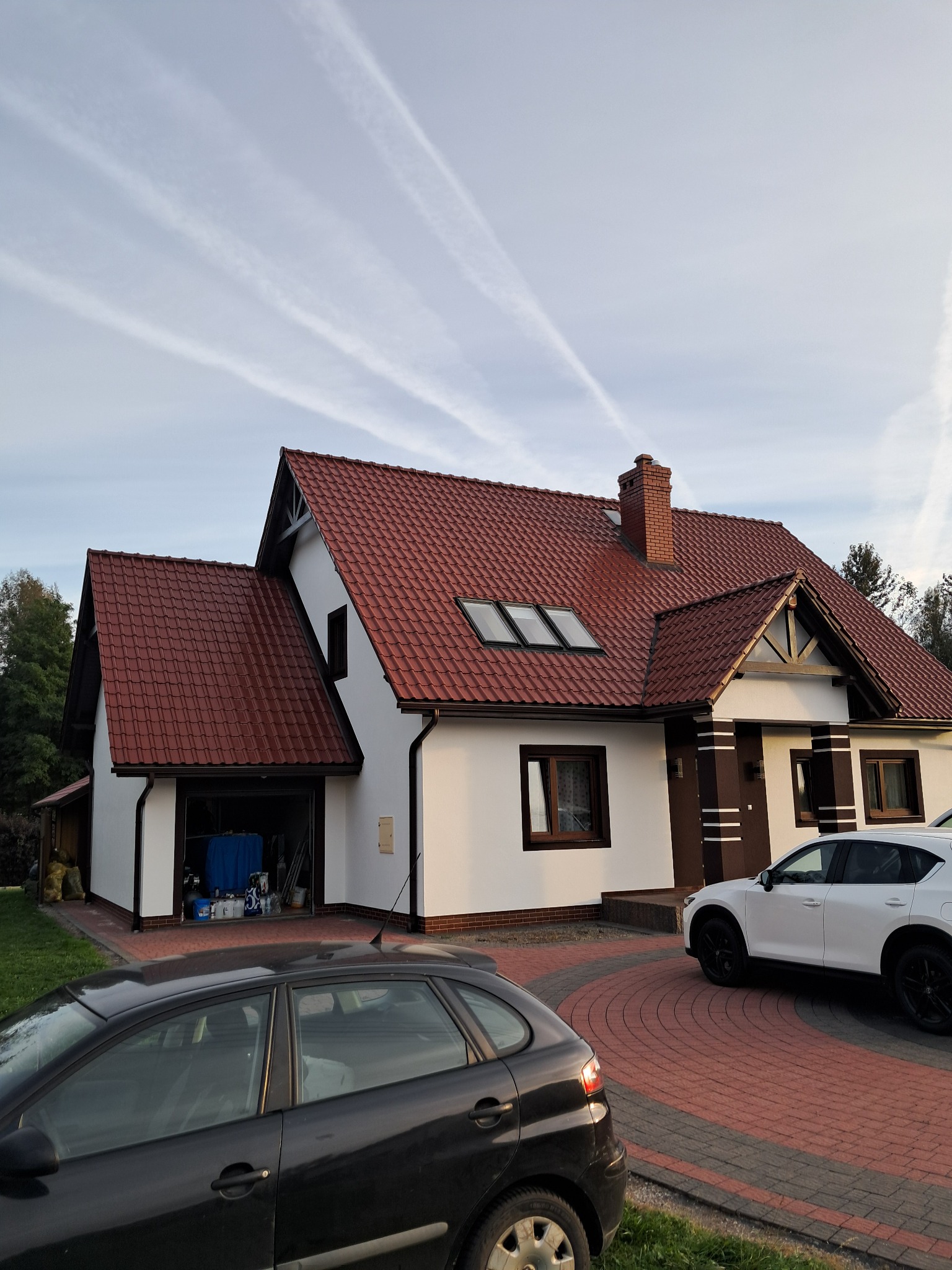 Biały dom jednorodzinny z czerwoną dachówką, widok z zewnątrz, z dwoma samochodami na podjeździe z kostki. Widoczne detale wykończenia elewacji i stolarki okiennej.