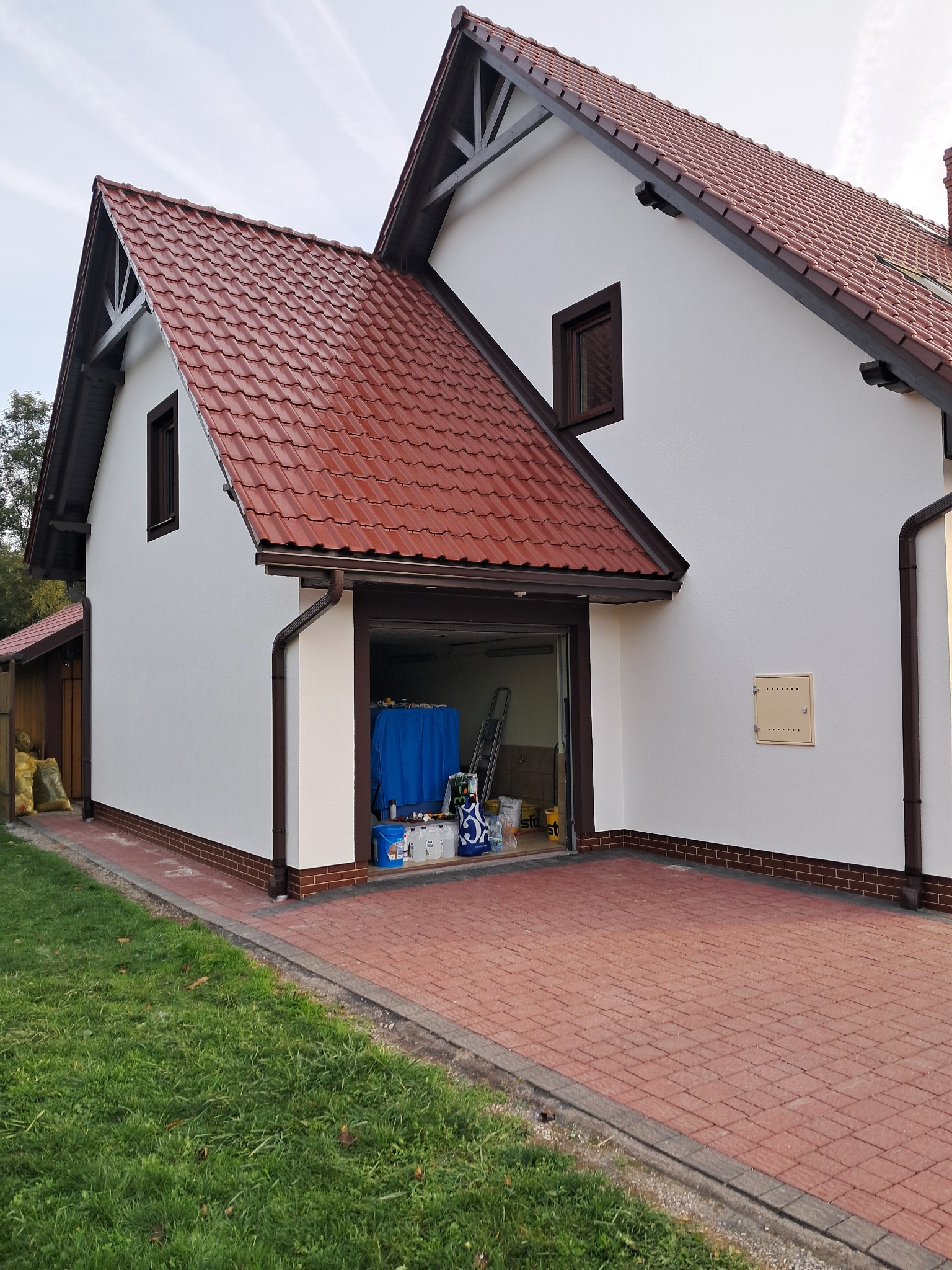 Biała elewacja domu z brązowymi detalami, widok na otwarty garaż z narzędziami malarskimi. Czerwona dachówka i kostka brukowa. Świeżo pomalowana ściana.