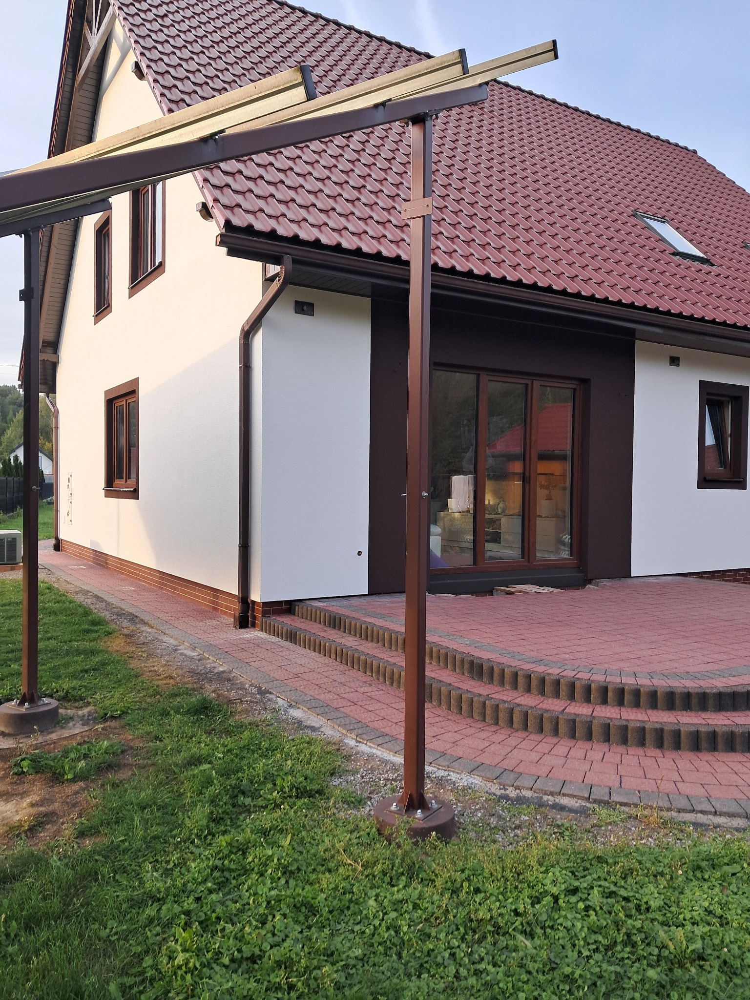 Dwukondygnacyjny dom z jasną elewacją i brązowymi akcentami, widoczny z boku, z fragmentem tarasu z czerwonej kostki i elementami konstrukcji zadaszenia.