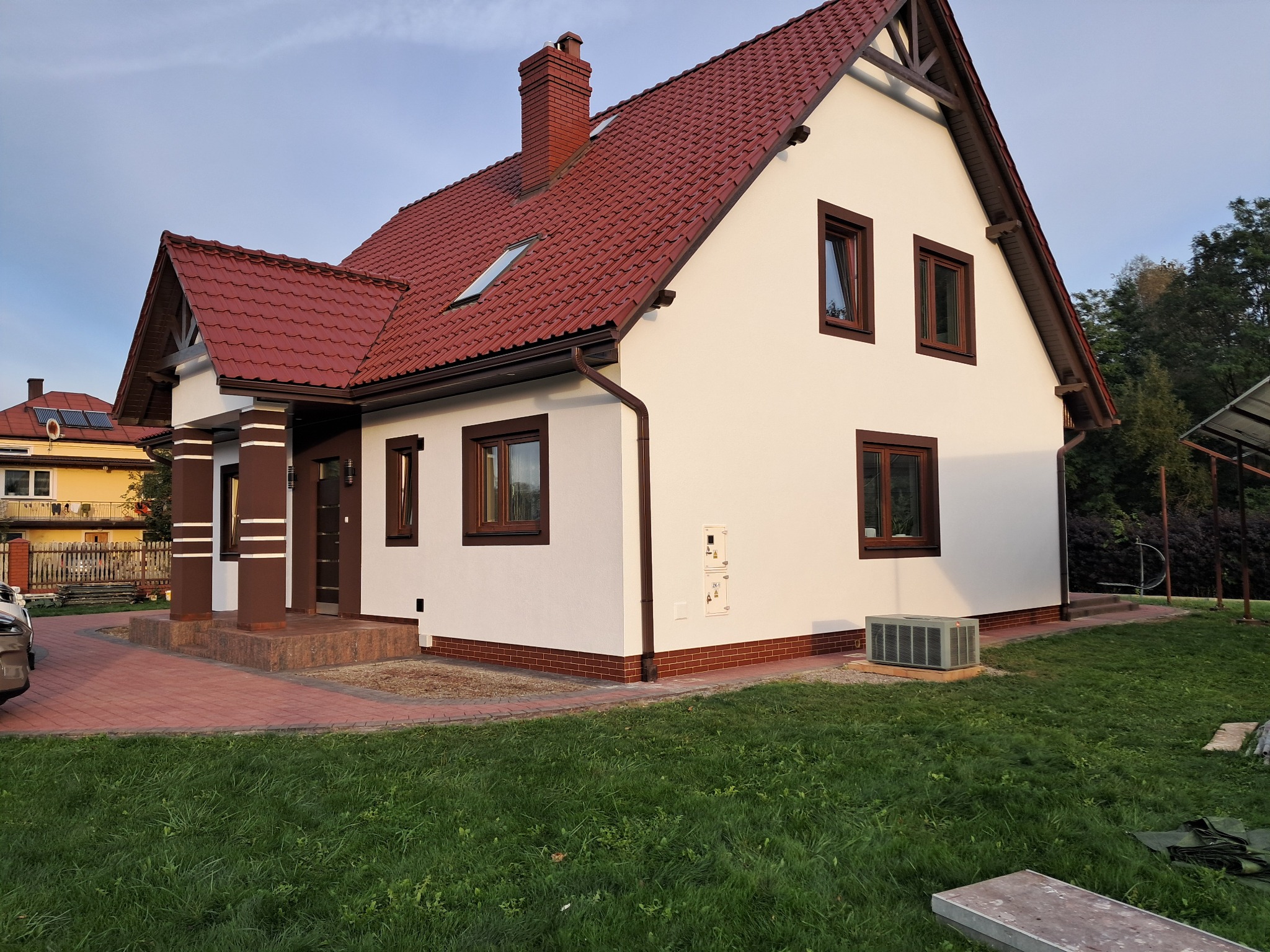 Biały dom jednorodzinny z czerwoną dachówką, brązowymi oknami i kolumnami werandy. Widoczne elementy wykończeniowe elewacji i zadbany trawnik.