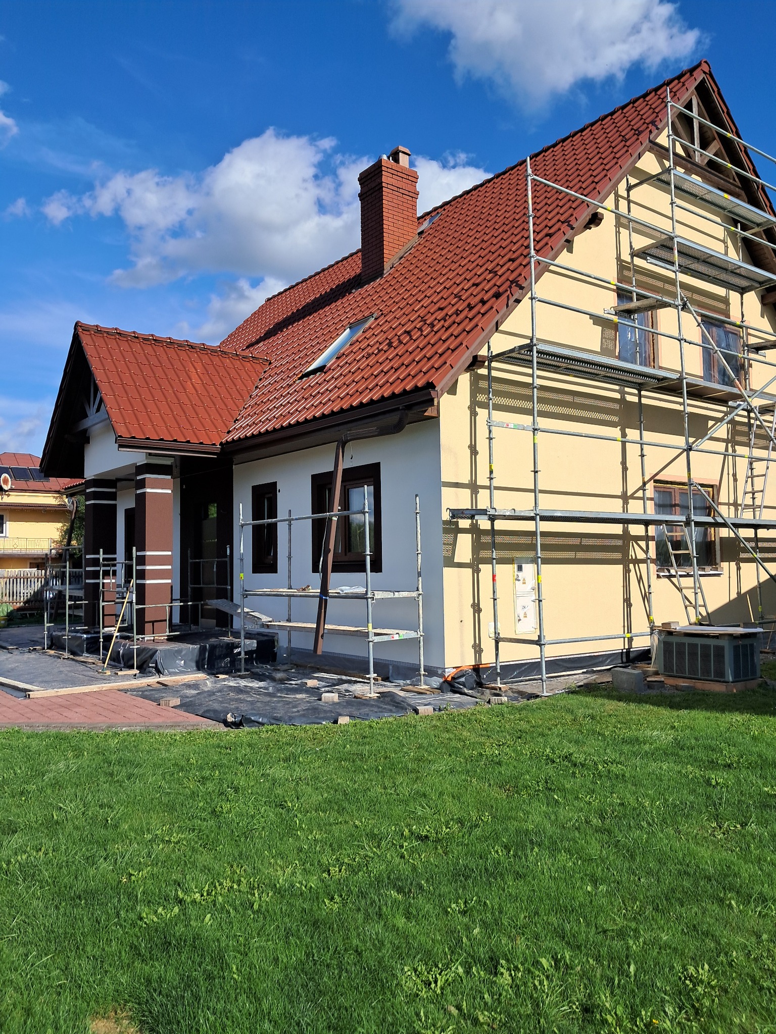 Dom jednorodzinny w Jarosławiu w trakcie prac elewacyjnych. Widoczne rusztowania, przygotowane podłoże i fragment pomalowanej ściany. Czerwona dachówka i błękitne niebo.