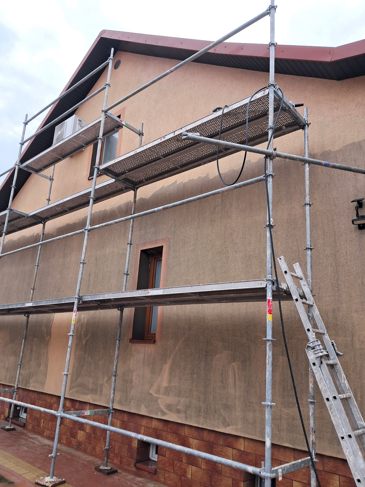 Dom w trakcie prac elewacyjnych: rusztowanie przy ścianie z widocznymi śladami po myciu. Fragment dachu z brązową dachówką. Widoczne okna i klimatyzator.