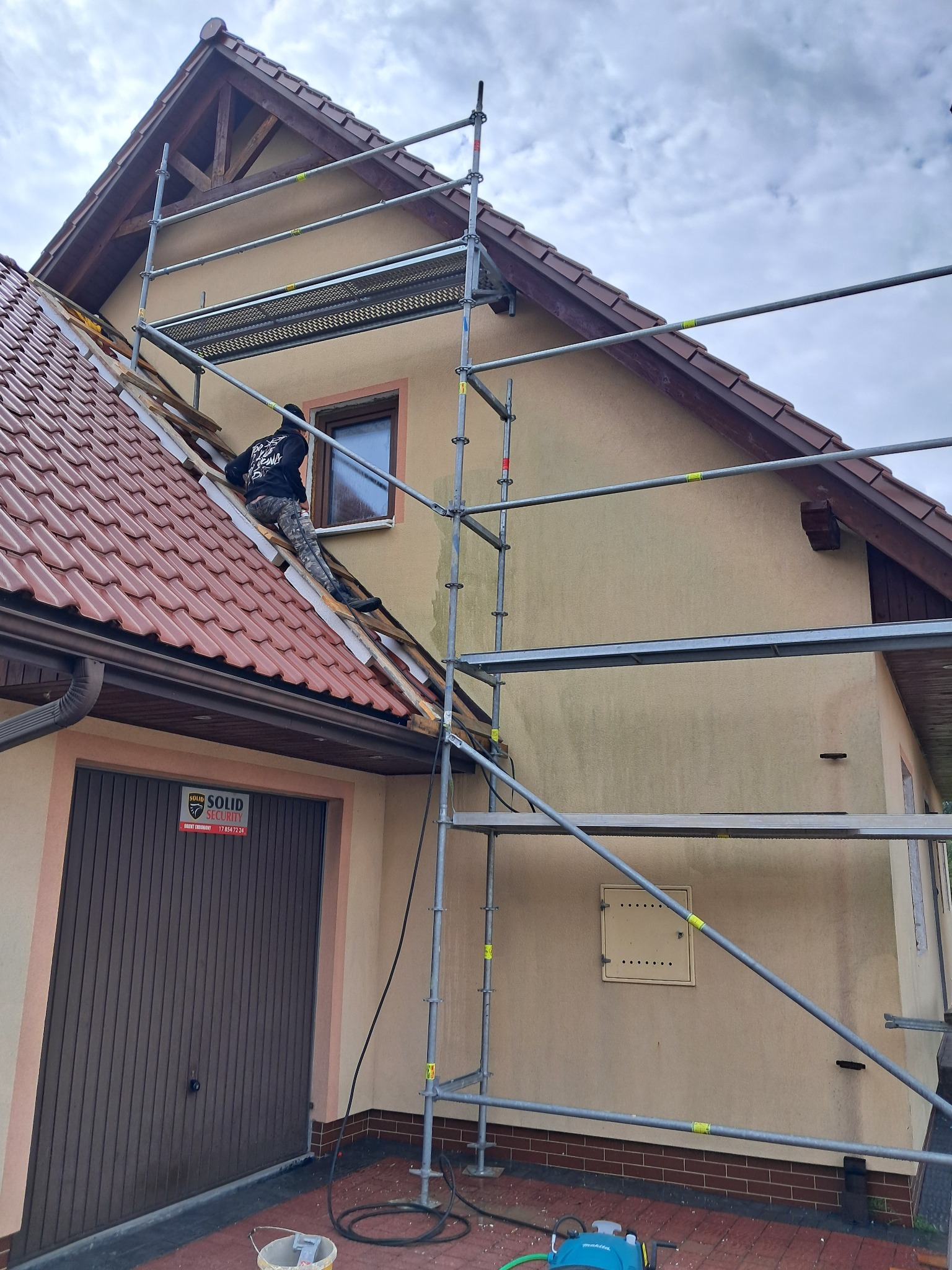 Pracownik na rusztowaniu przy domu z częściowo zdjętym dachem, przygotowuje elewację do prac. Widoczne narzędzia i materiały budowlane.