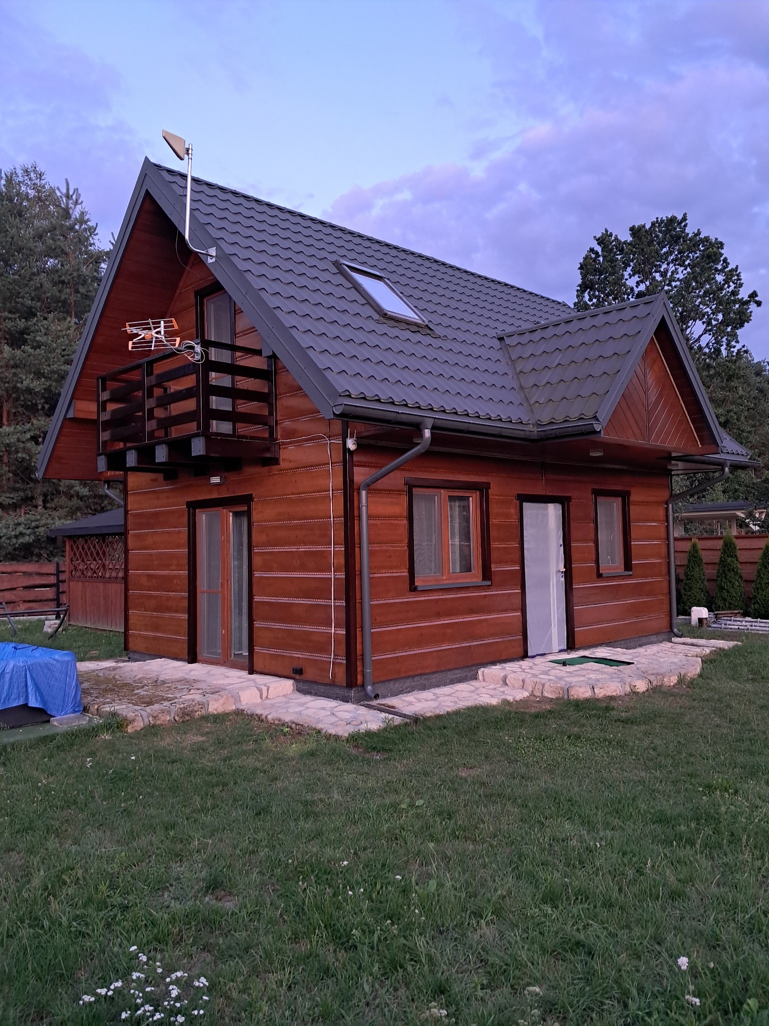 Drewniany dom z balkonem i szarą dachówką, oświetlony wieczornym światłem. Widoczne detale wykończenia elewacji i otoczenie domu z zielonym trawnikiem.