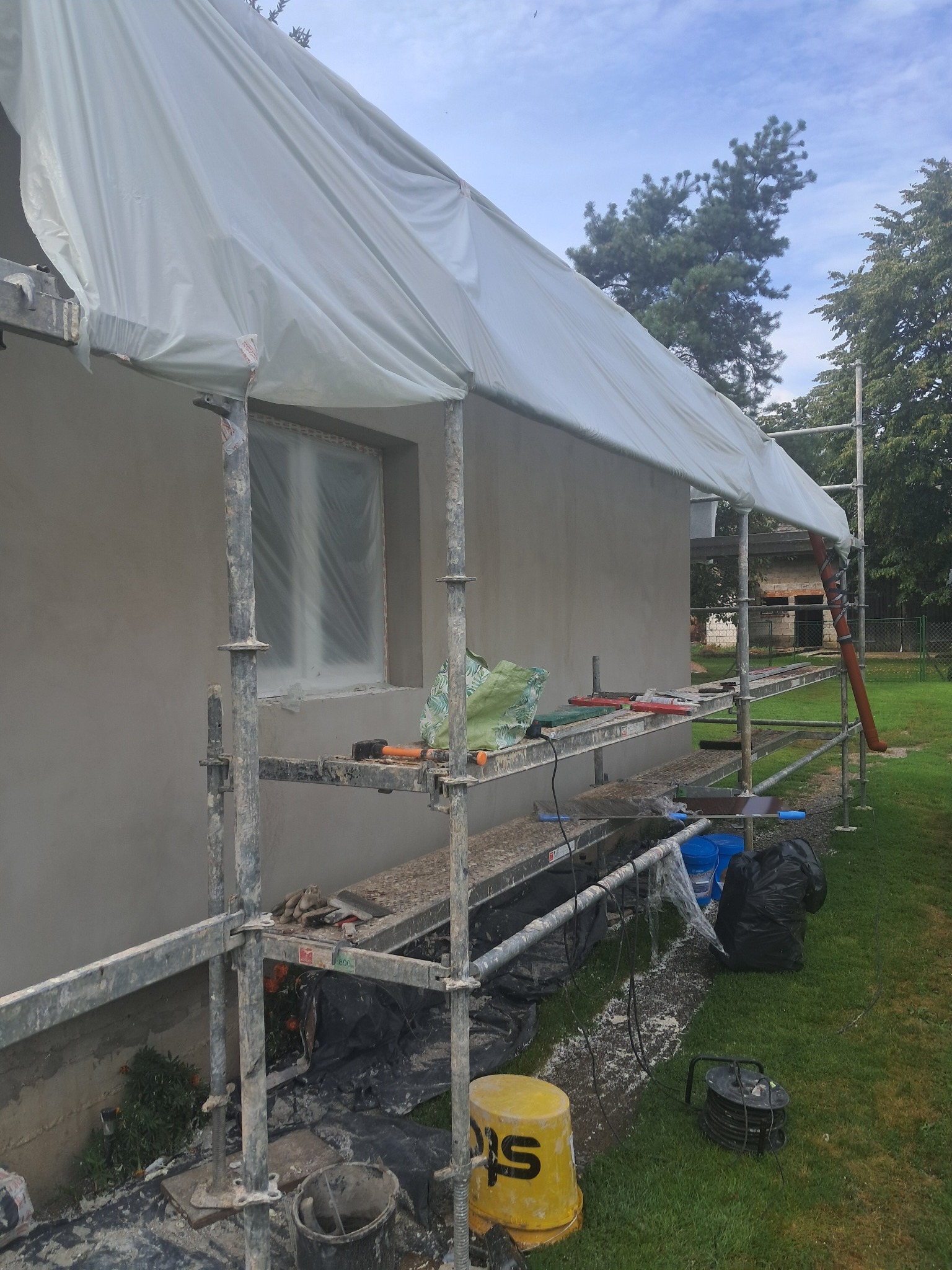 Świeżo otynkowana elewacja domu jednorodzinnego, zabezpieczona folią i rusztowaniem. Widoczne narzędzia pracy i wiadro z farbą. Tło: zieleń drzew i błękitne niebo.