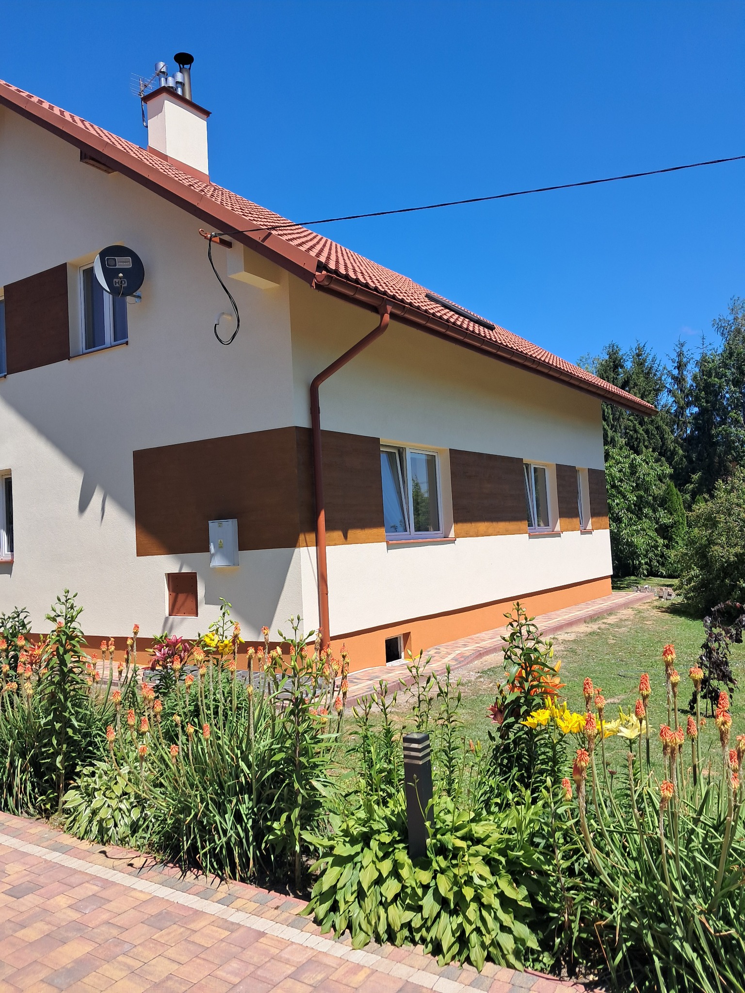 Elewacja domu jednorodzinnego po renowacji, z dekoracyjnymi panelami imitującymi drewno, otoczona kwitnącym ogrodem i kostką brukową. Słoneczny dzień.