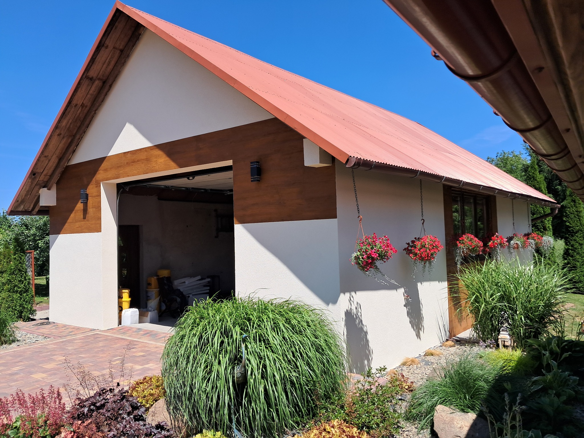 Wykończony budynek gospodarczy z otwartymi drzwiami garażowymi, pomalowany na biało z brązowymi akcentami, czerwonym dachem i wiszącymi kwiatami.